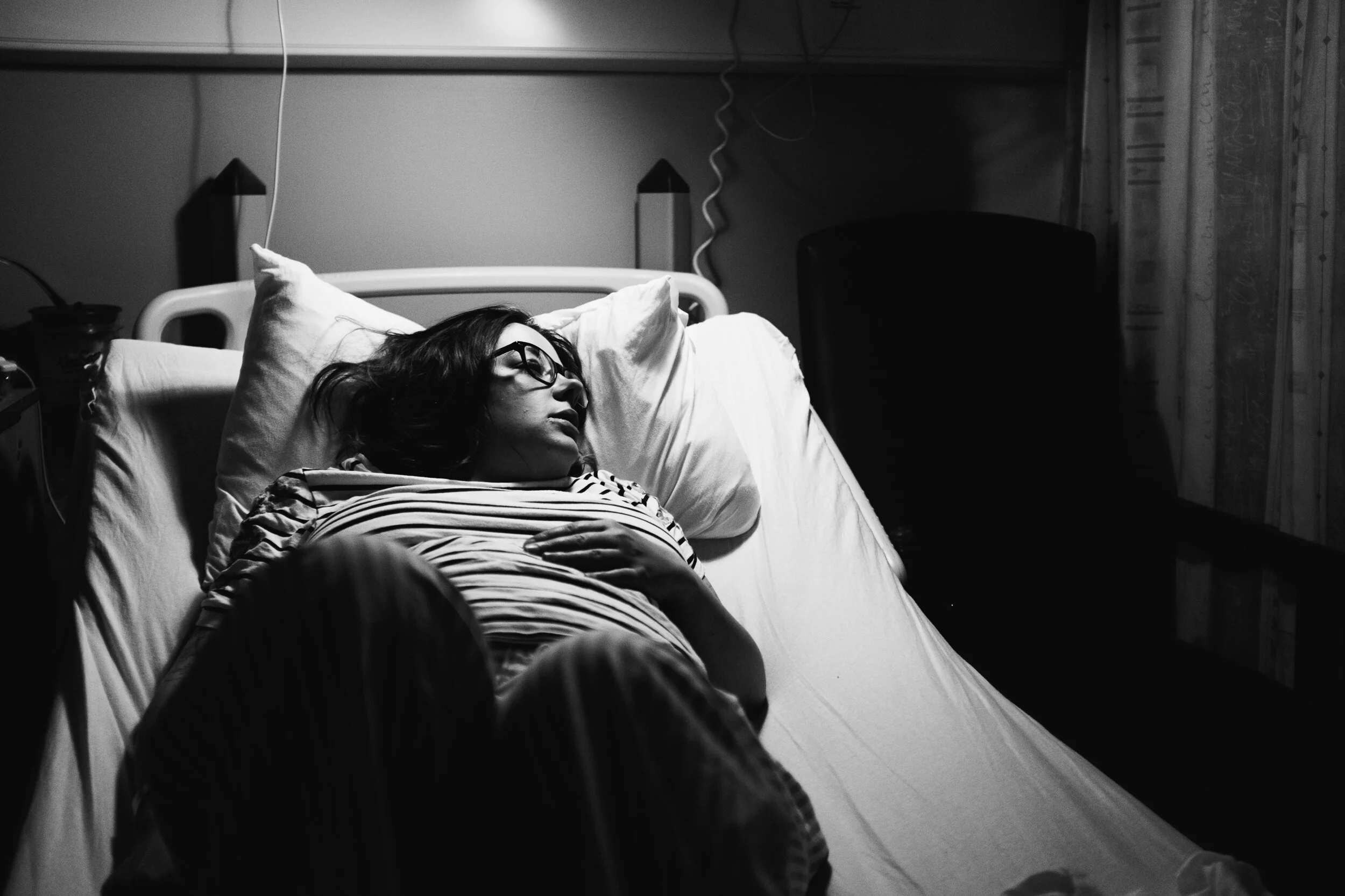 A woman with glasses and dark hair lying in a hospital bed with her eyes closed, covered with a striped blanket, in a hospital room with medical equipment and supplies around.