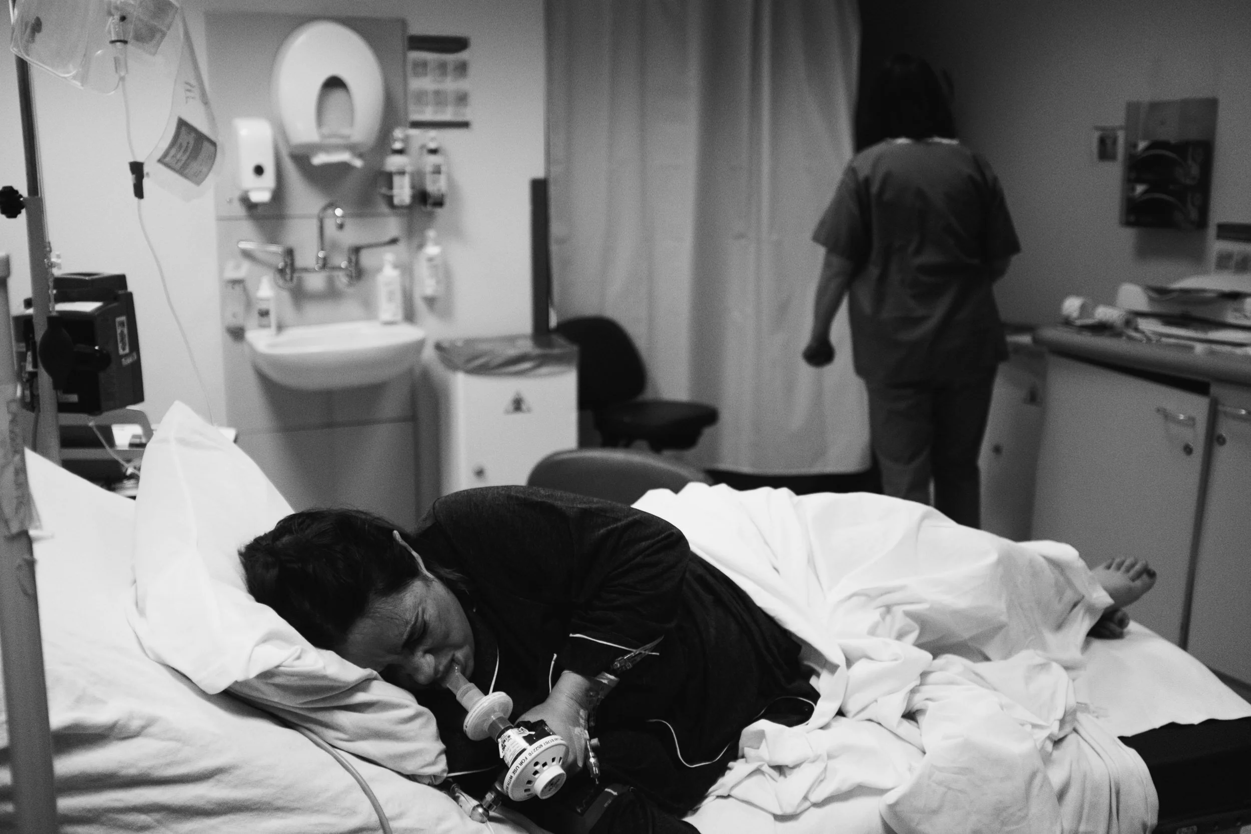 A patient lying in a hospital bed with a breathing tube, receiving medical care. A person, likely a medical professional, is walking away in the background in a hospital room with medical equipment and supplies.