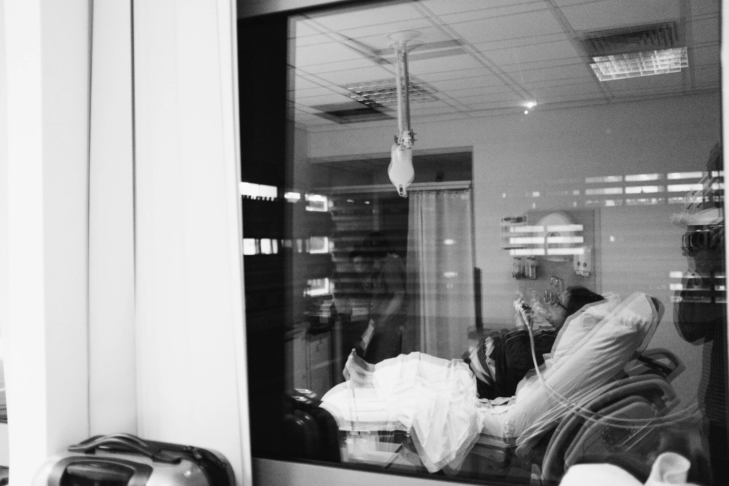 A black and white photo shows a person in a hospital bed with medical equipment around, including an IV drip and various medical devices, seen through a glass window or mirror.