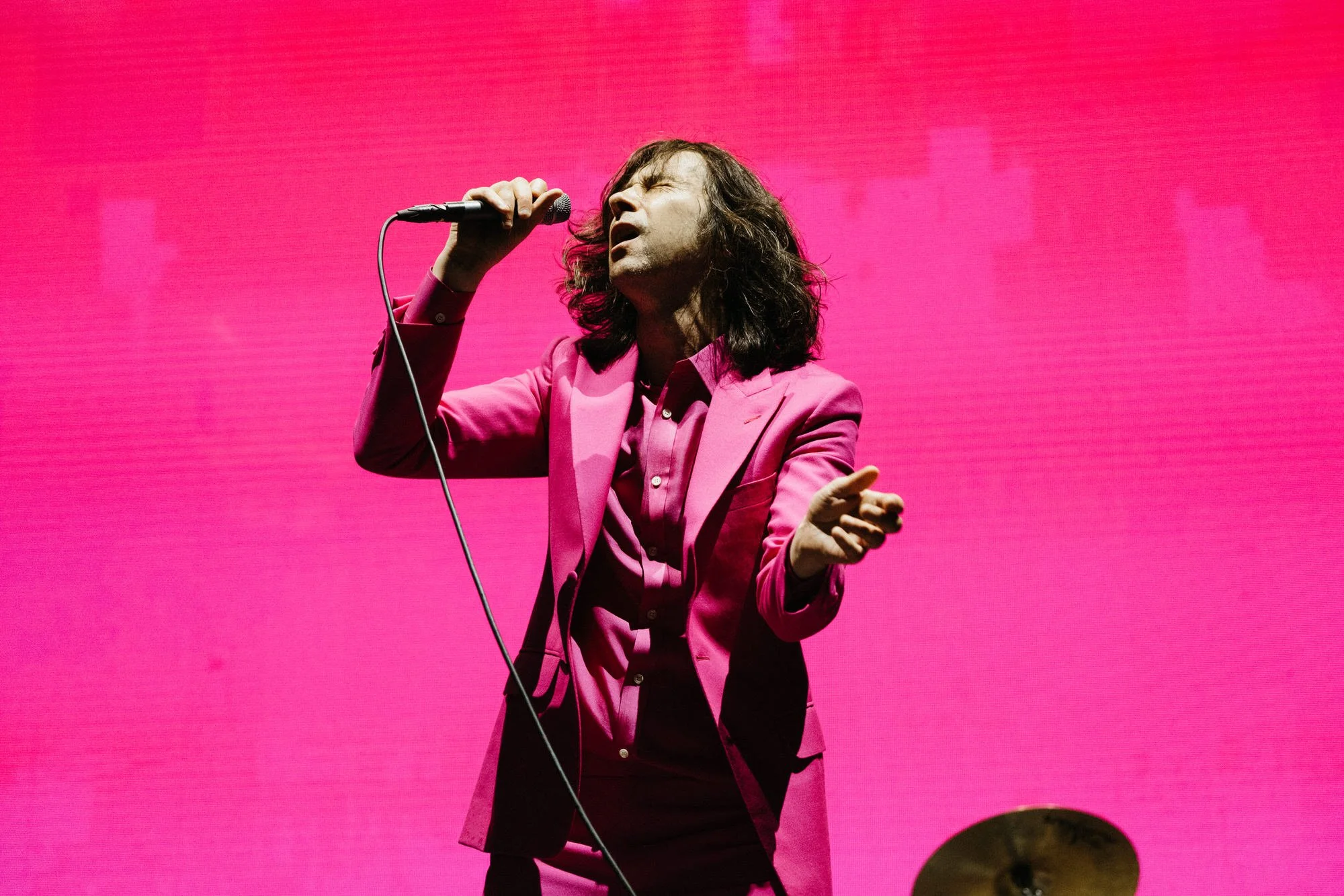 A man in a pink suit and shirt singing passionately into a microphone against a pink background.
