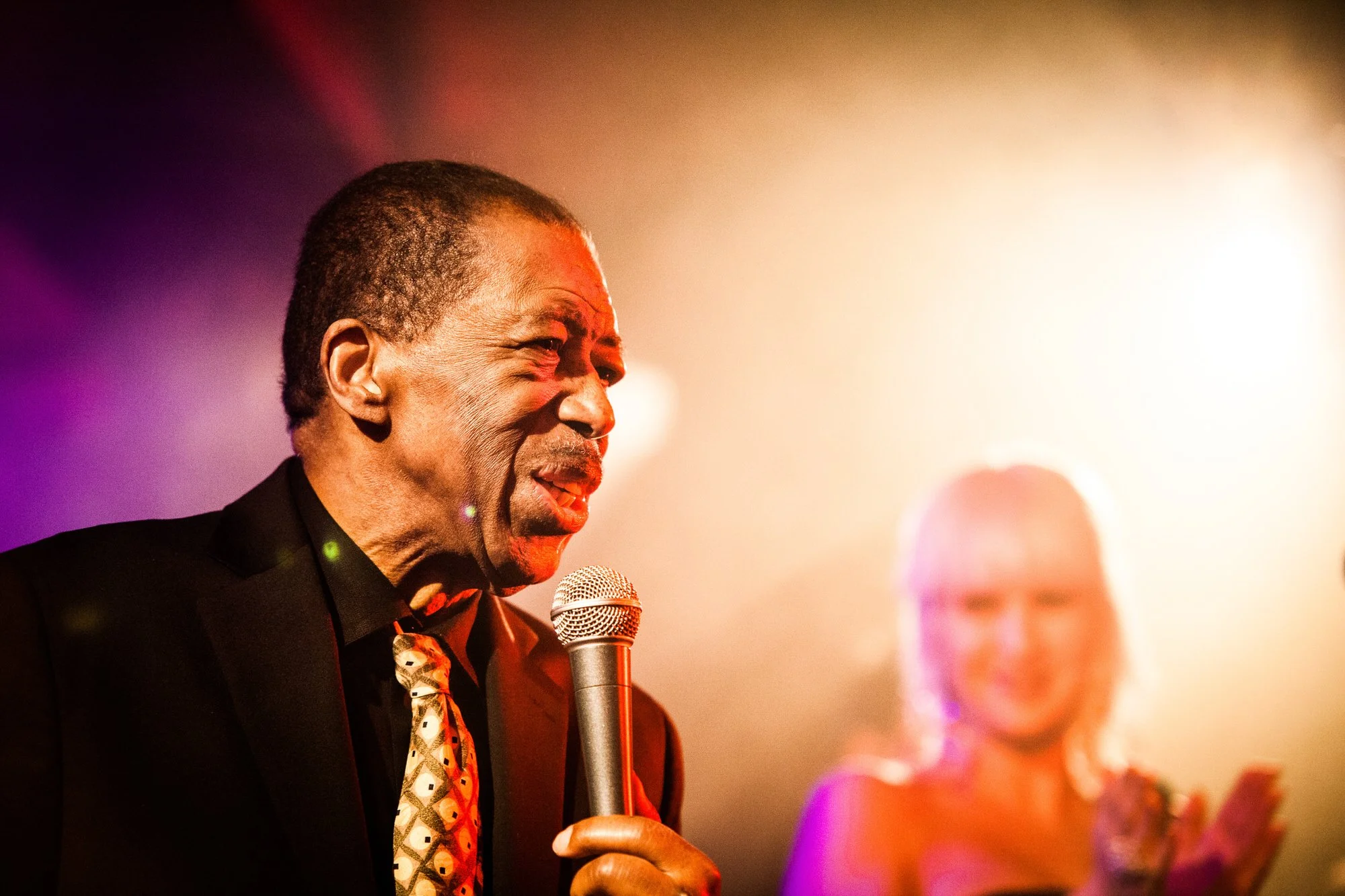 An elderly man with short gray hair, dressed in a black suit, holding a microphone, speaking or singing at an event. The background is blurred with multicolored lights and a woman in glasses is visible behind him.