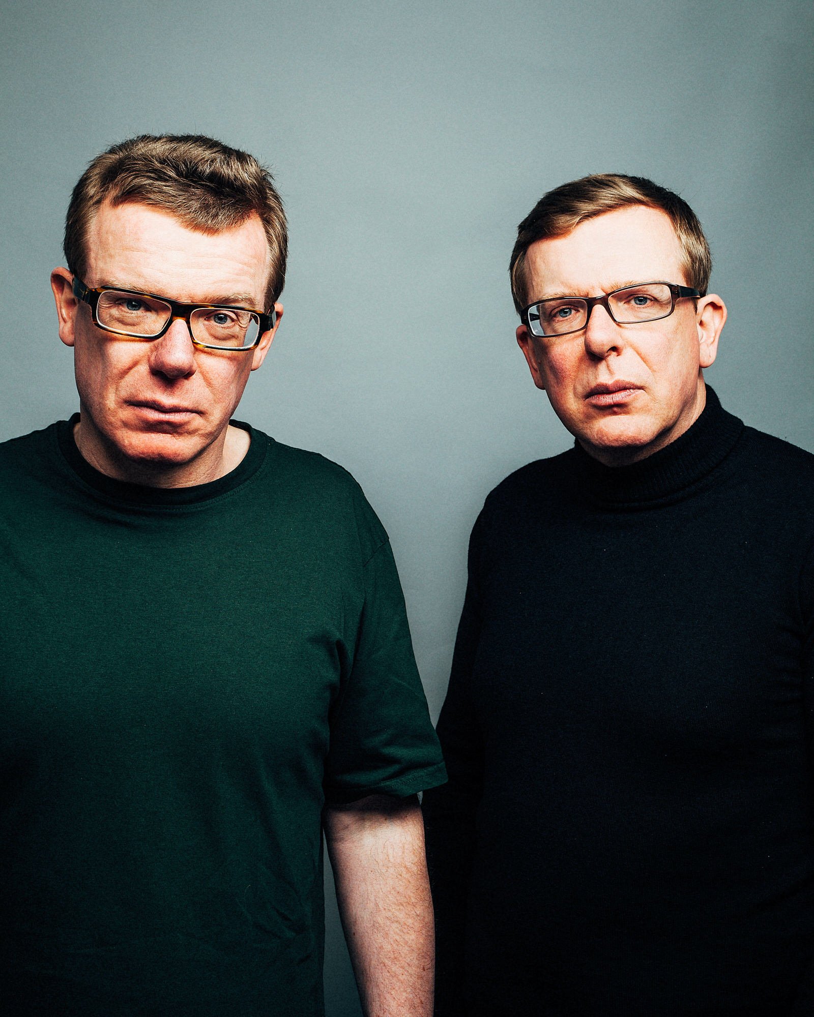 Close-up of two men with glasses facing forward, partially visible in a gray background.