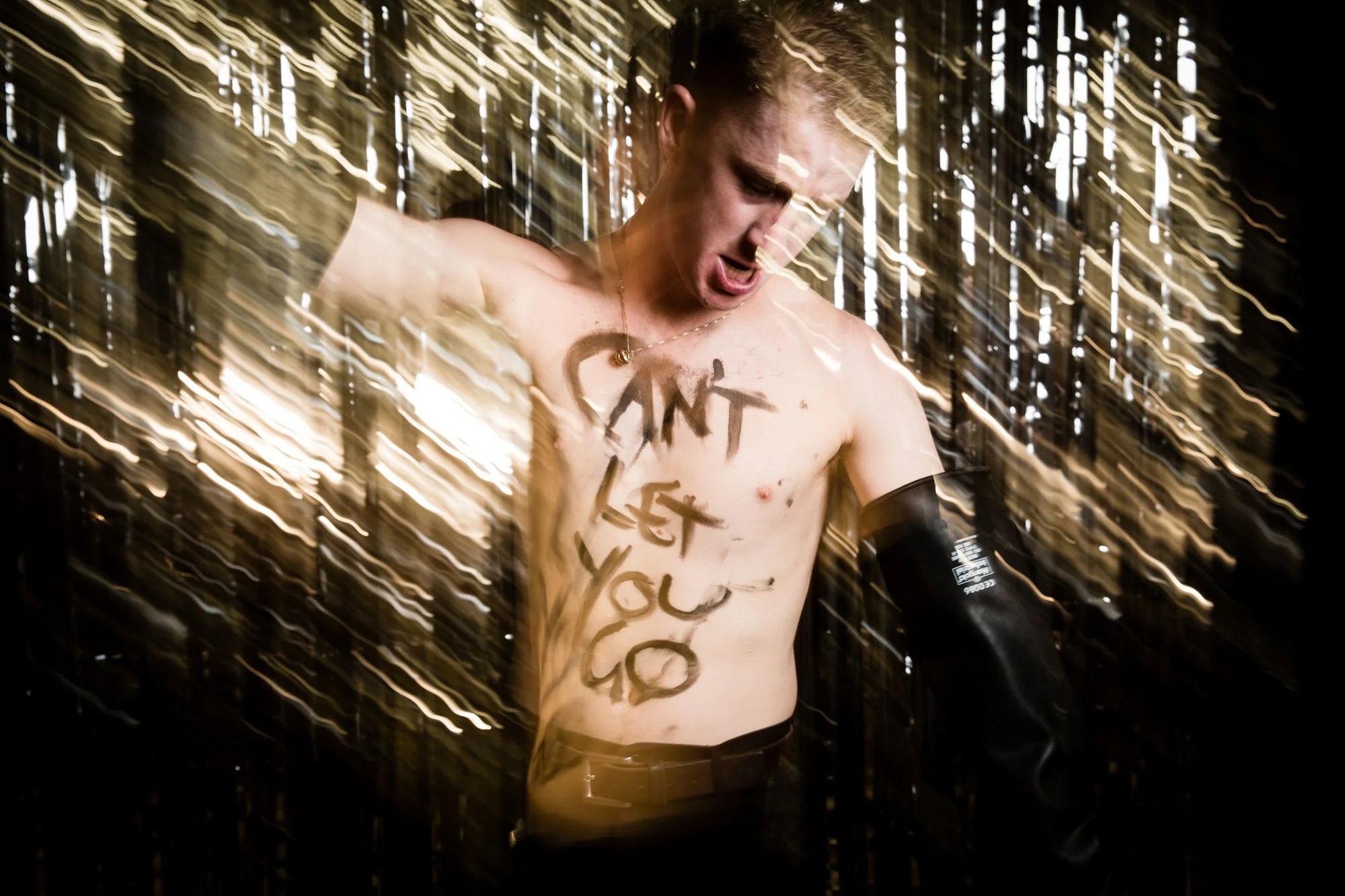 A shirtless young man with graffiti writing on his chest, shouting with a panicked expression, surrounded by streaks of light in motion, in a dark setting.