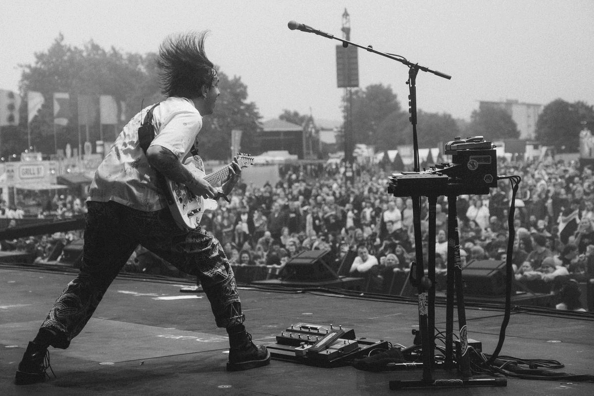 Musician on stage playing guitar with an energetic stance, microphone stand and equipment setup, audience in background at outdoor event.