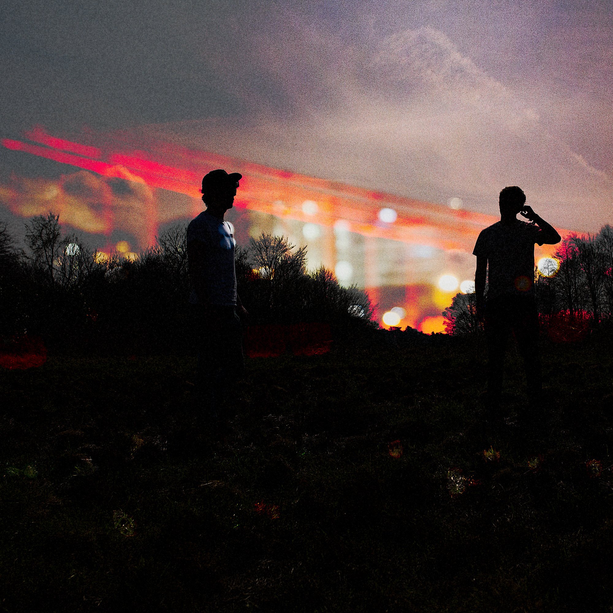 Silhouettes of two people standing outdoors during sunset, with leafless trees and colorful sky with clouds and bokeh lights in the background.