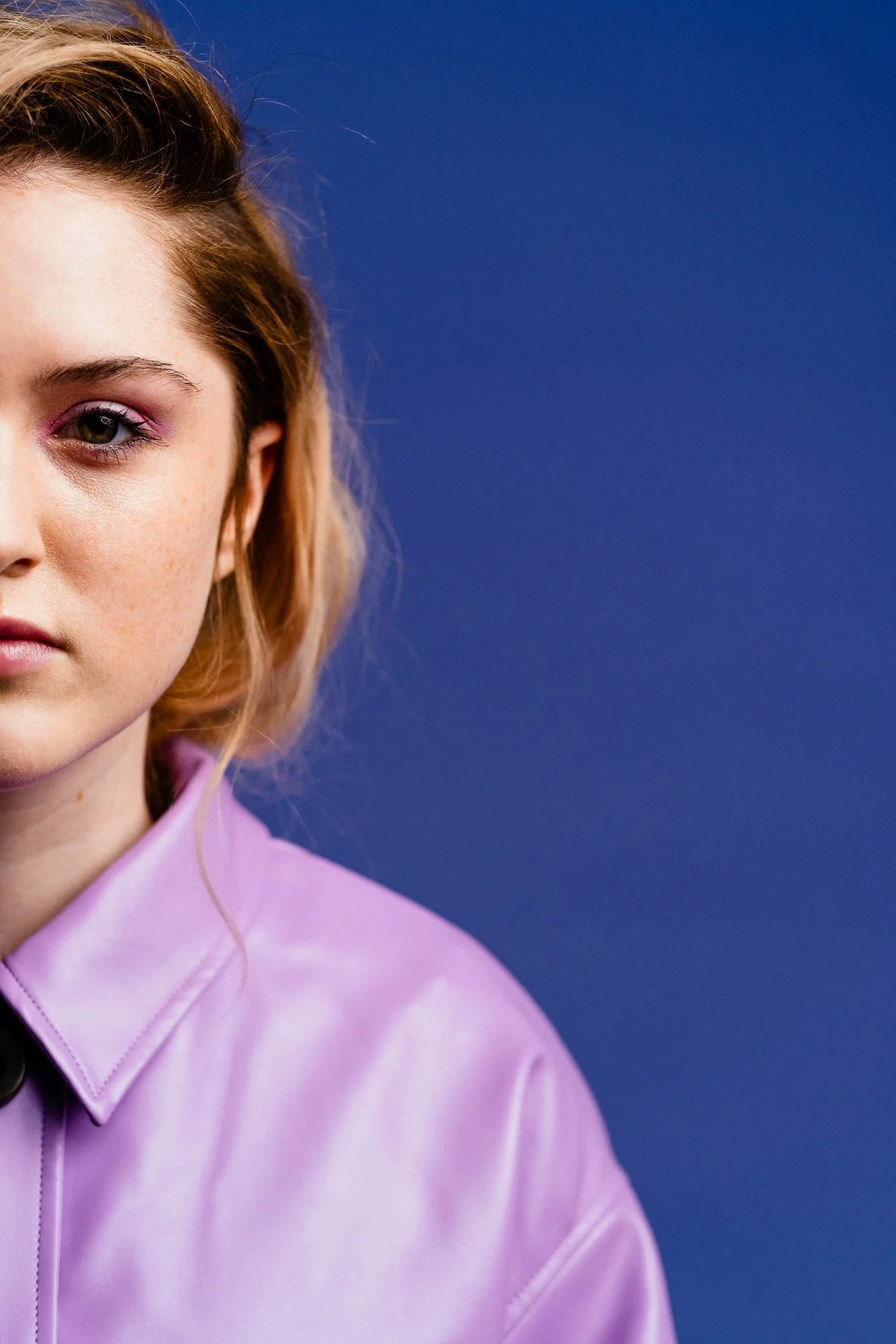 Partial view of a woman with light skin, red hair, wearing purple lipstick and a lavender jacket, against a solid blue background.