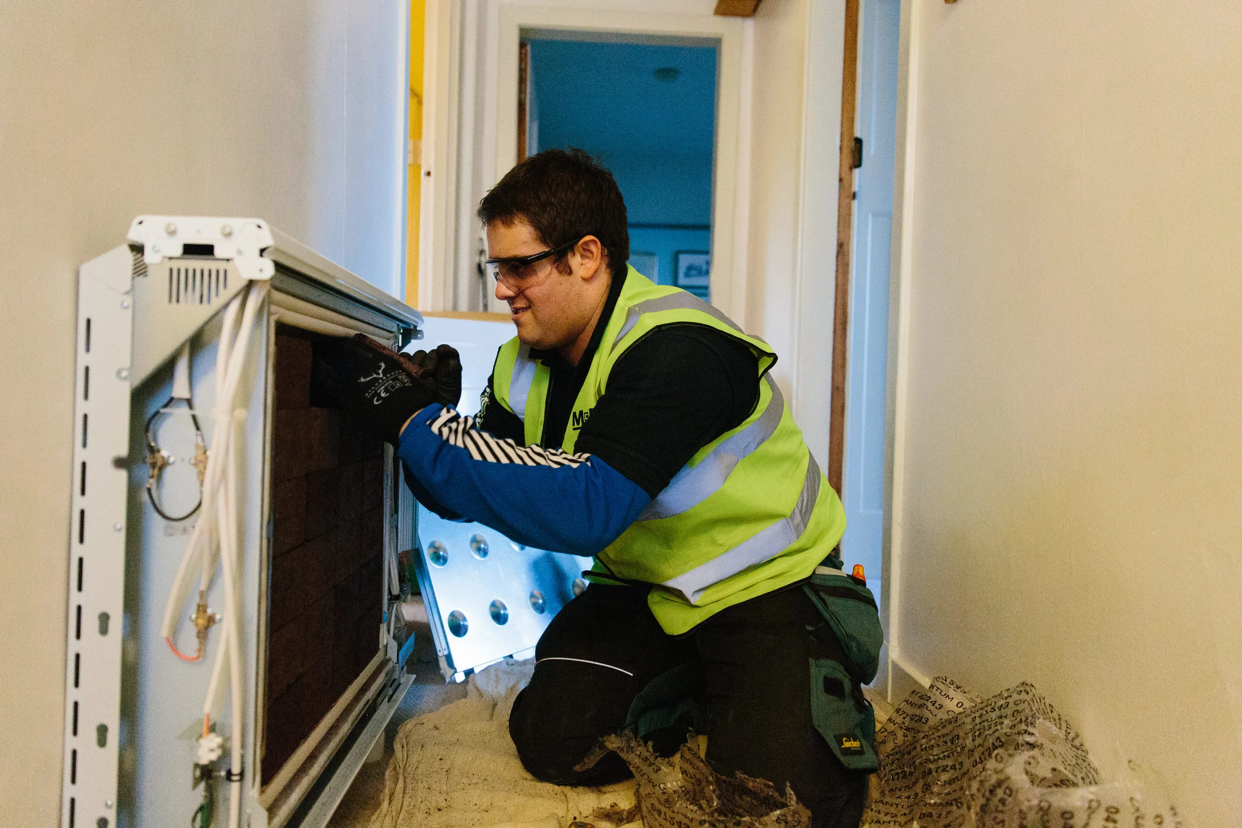A technician wearing safety goggles, a yellow high-visibility vest, and gloves is kneeling inside a narrow hallway, working on an electrical or network panel. There are wires and equipment within the panel, and the background shows the hallway leadin