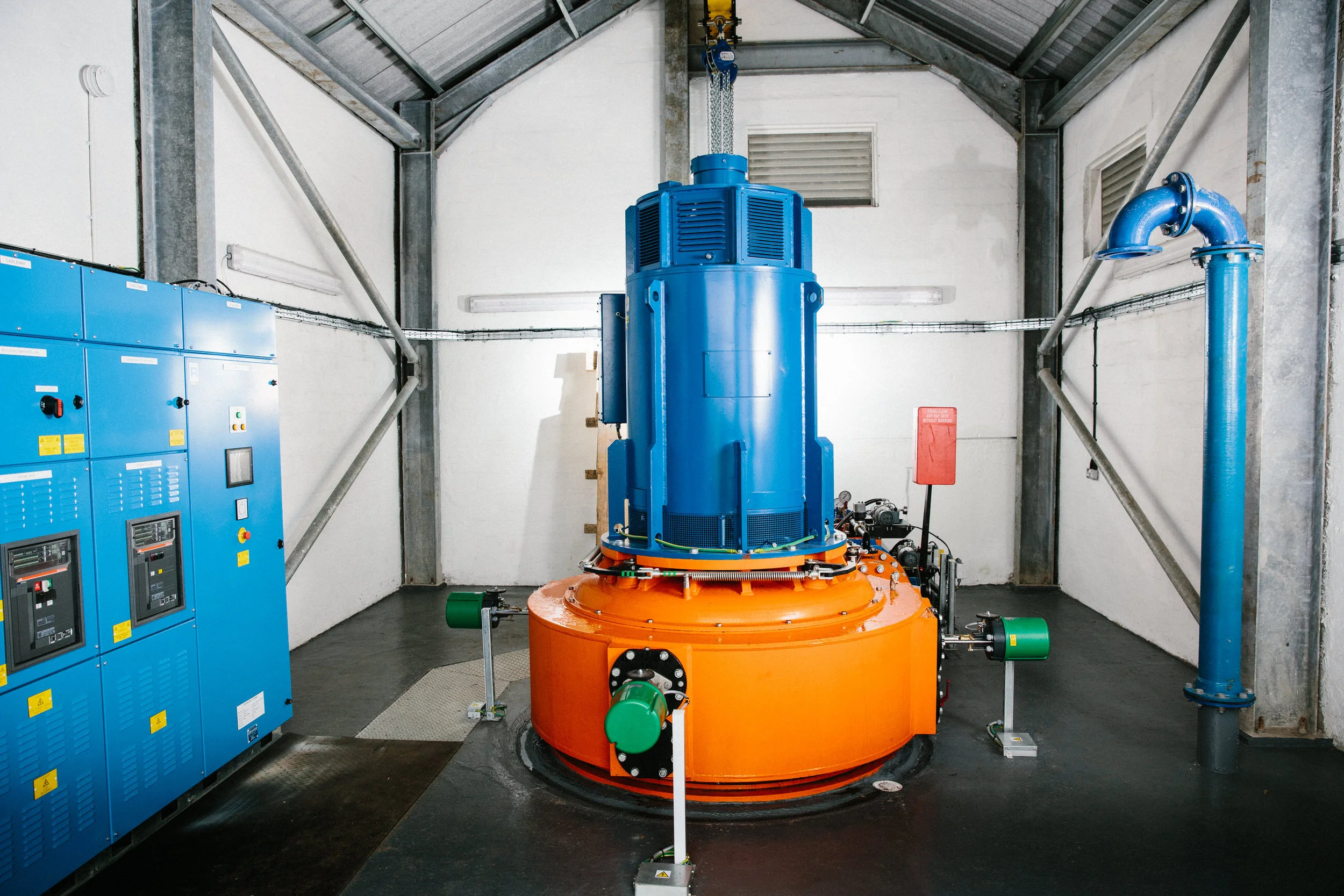 Industrial machinery inside a facility with a tall blue and orange equipment piece, blue electrical panels to the left, and blue piping on the right, all within a metal-framed building.