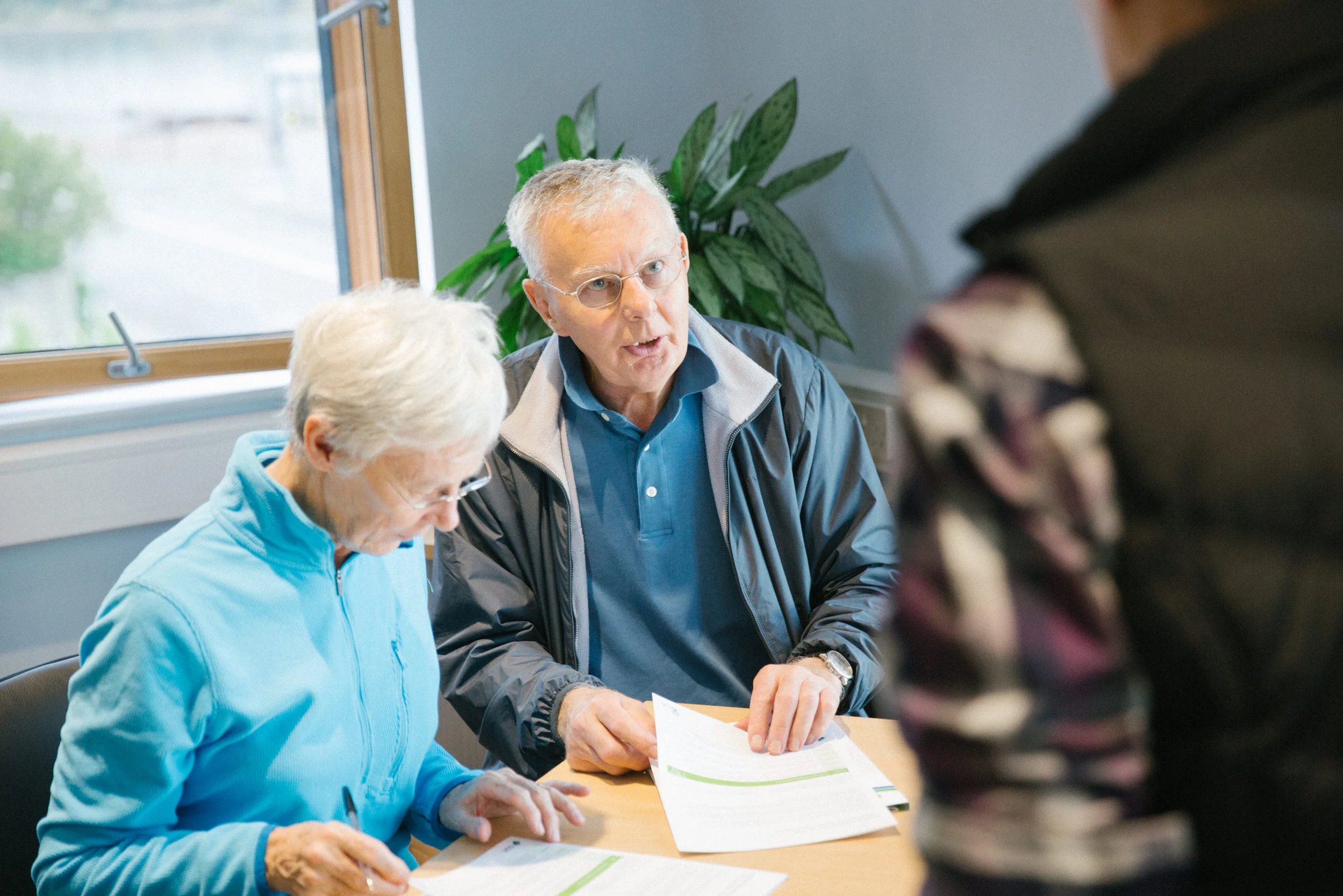 An elderly woman and a middle-aged man sitting at a table, looking at papers, while a person in the foreground is partially visible with a floral shirt. The elderly woman is wearing glasses and a blue jacket, the man has gray hair, glasses, and a blu