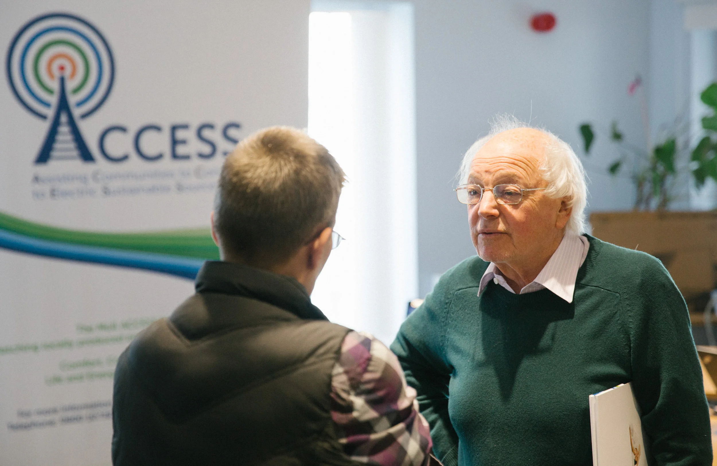 An elderly man with glasses and white hair talking to a younger person with short, light brown hair at an event with a banner that reads 'ACCESS' in the background.
