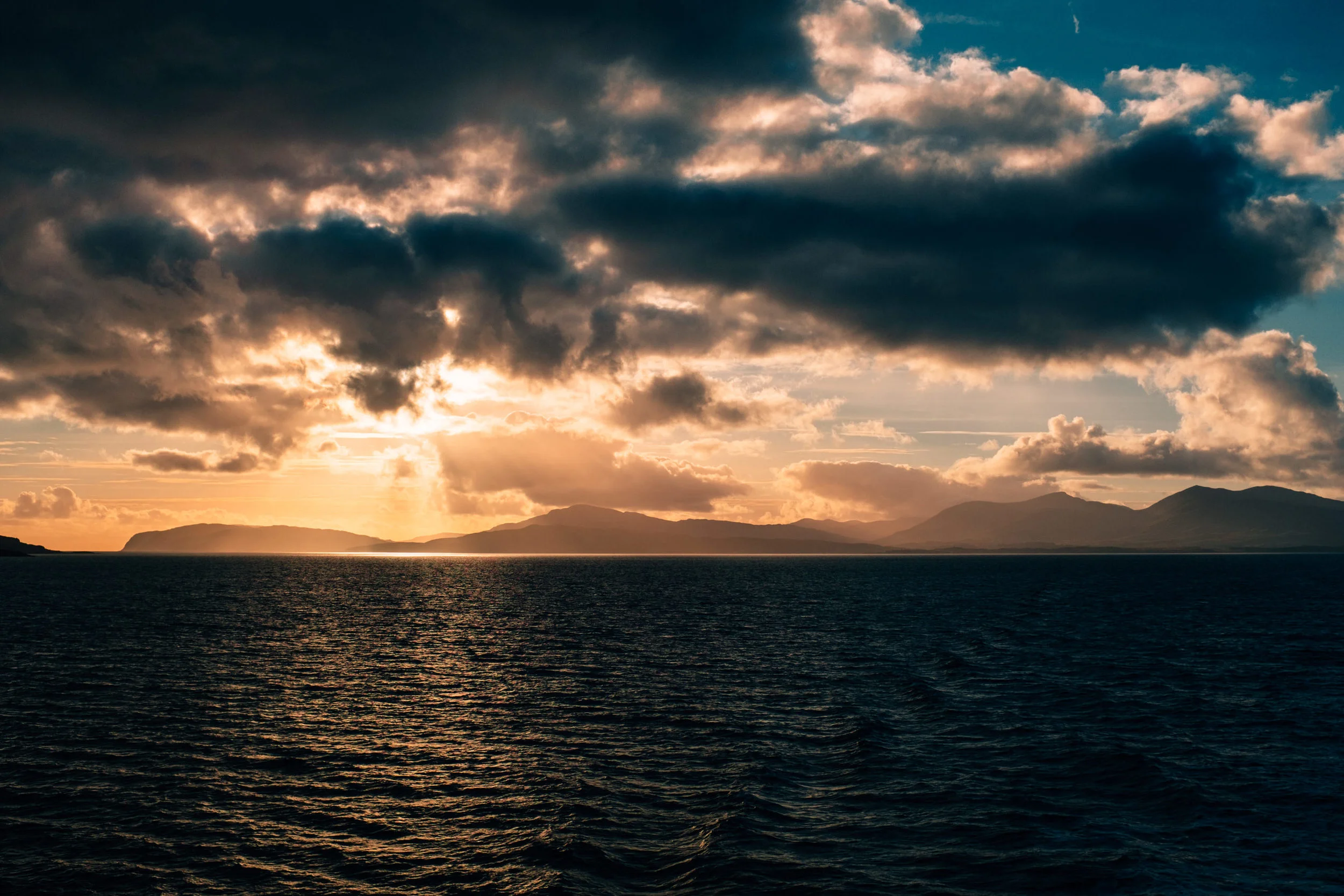 Sunset over a body of water with mountains in the distance and dark clouds in the sky.