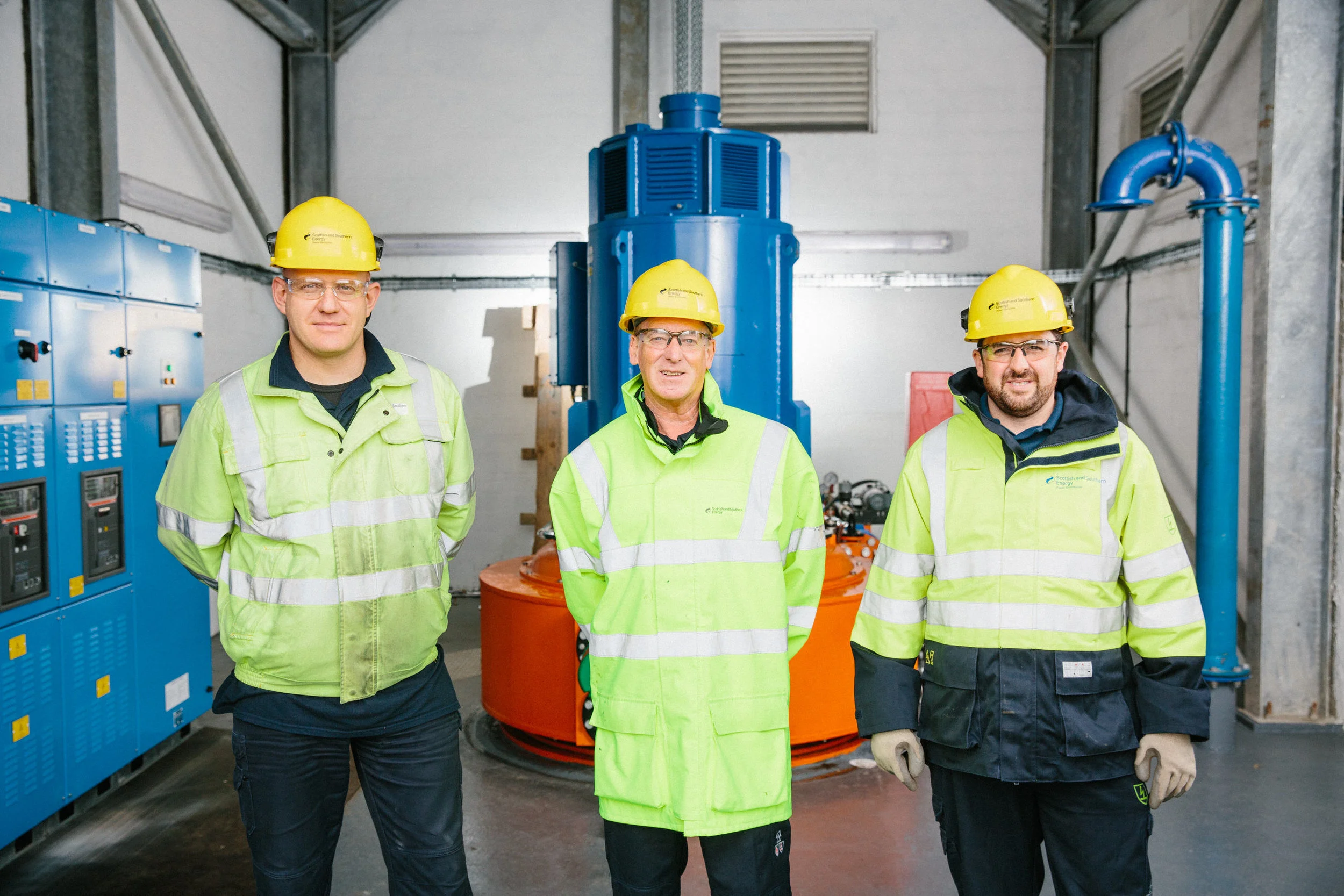 Three workers in high-visibility green jackets and yellow helmets standing inside an industrial facility with blue and orange machinery around.