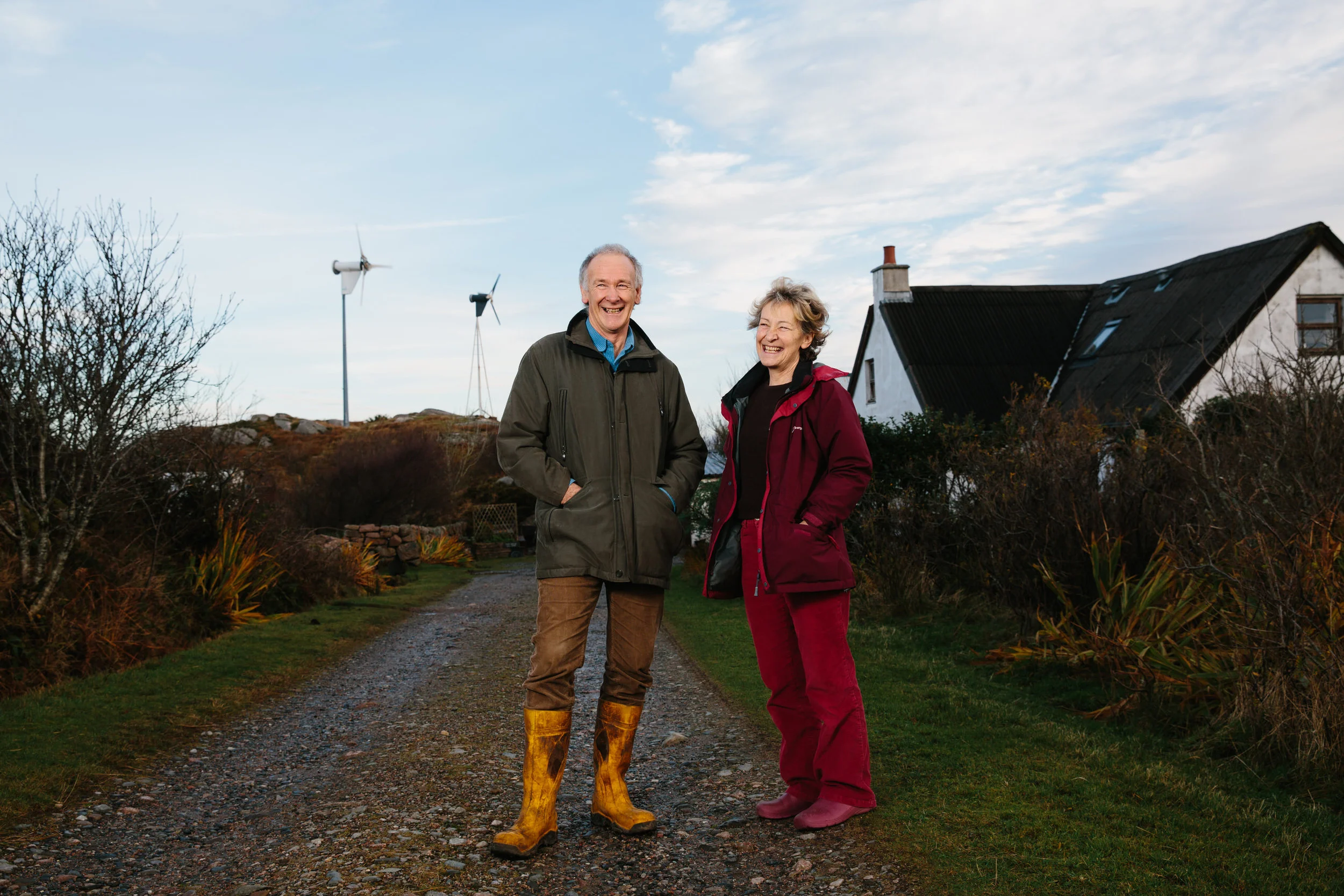 An older man and woman standing on a dirt path outdoors, smiling, with wind turbines and a house in the background, during daytime.