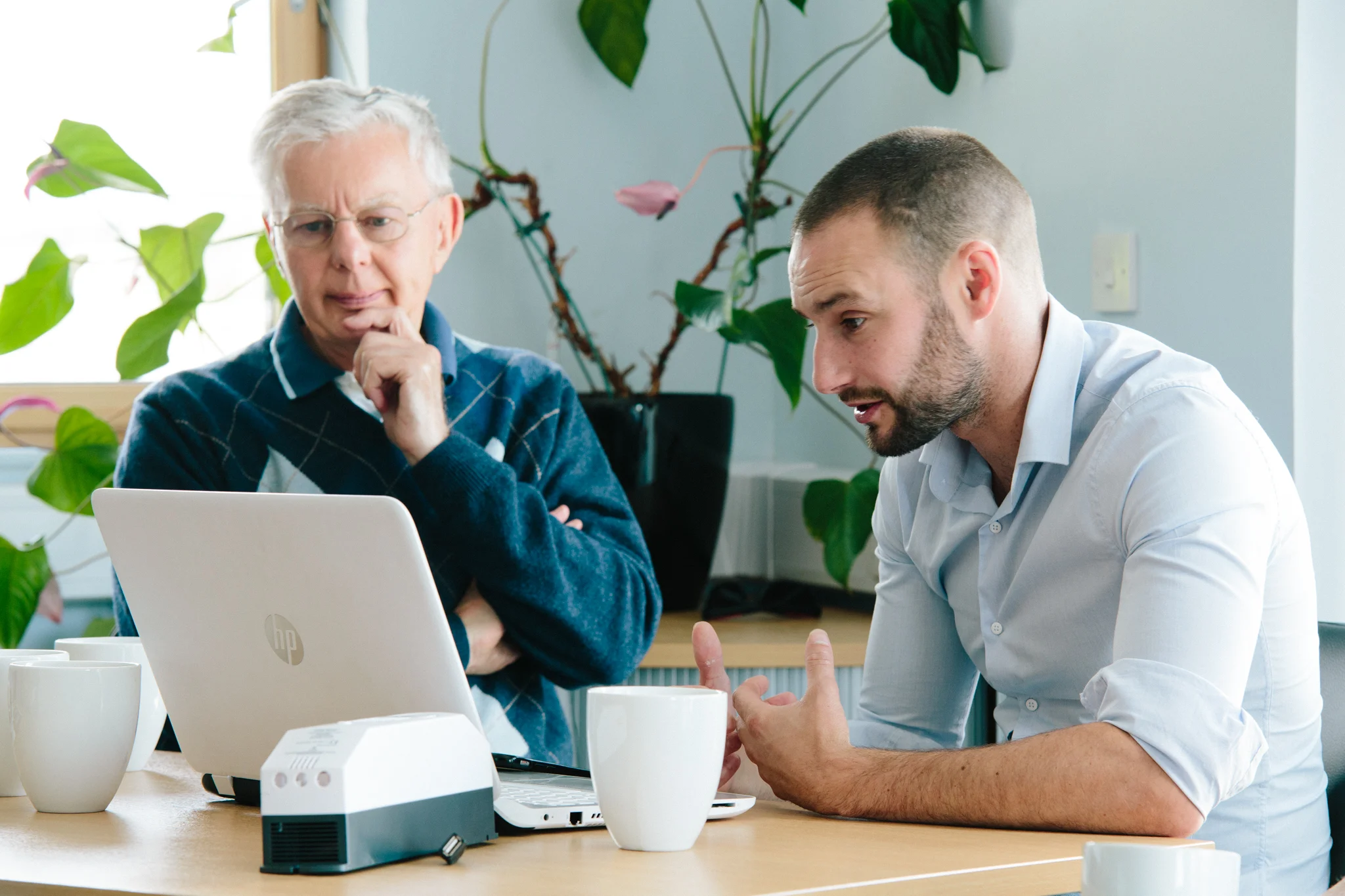 Two men are seated at a table working together with a laptop and other equipment in an office or meeting room. The older man with white hair and glasses looks thoughtful, while the younger man with a beard and short dark hair appears to be explaining