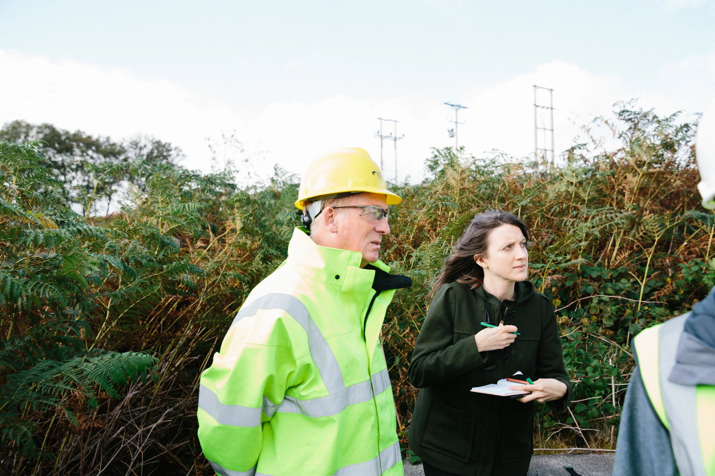 Two people are outdoors among dense greenery, one wearing a bright yellow safety helmet and high-visibility safety jacket, and the other holding a notebook and pen, engaged in a discussion.