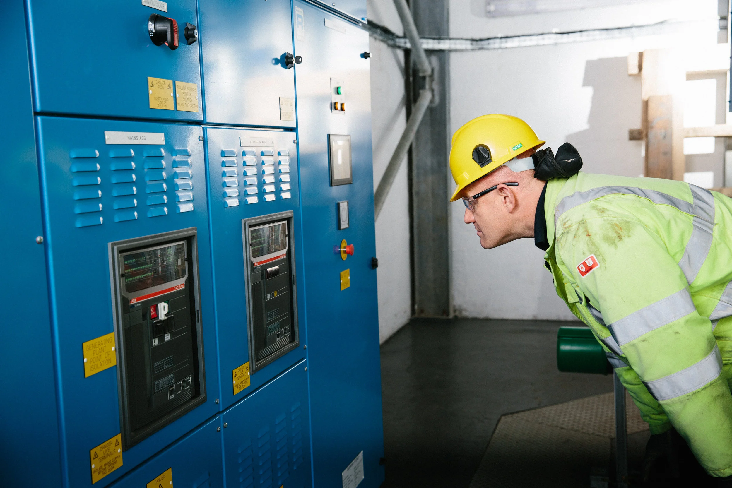 A worker wearing a yellow safety helmet, safety glasses, and a high-visibility green jacket is inspecting an electrical control panel in an industrial setting.