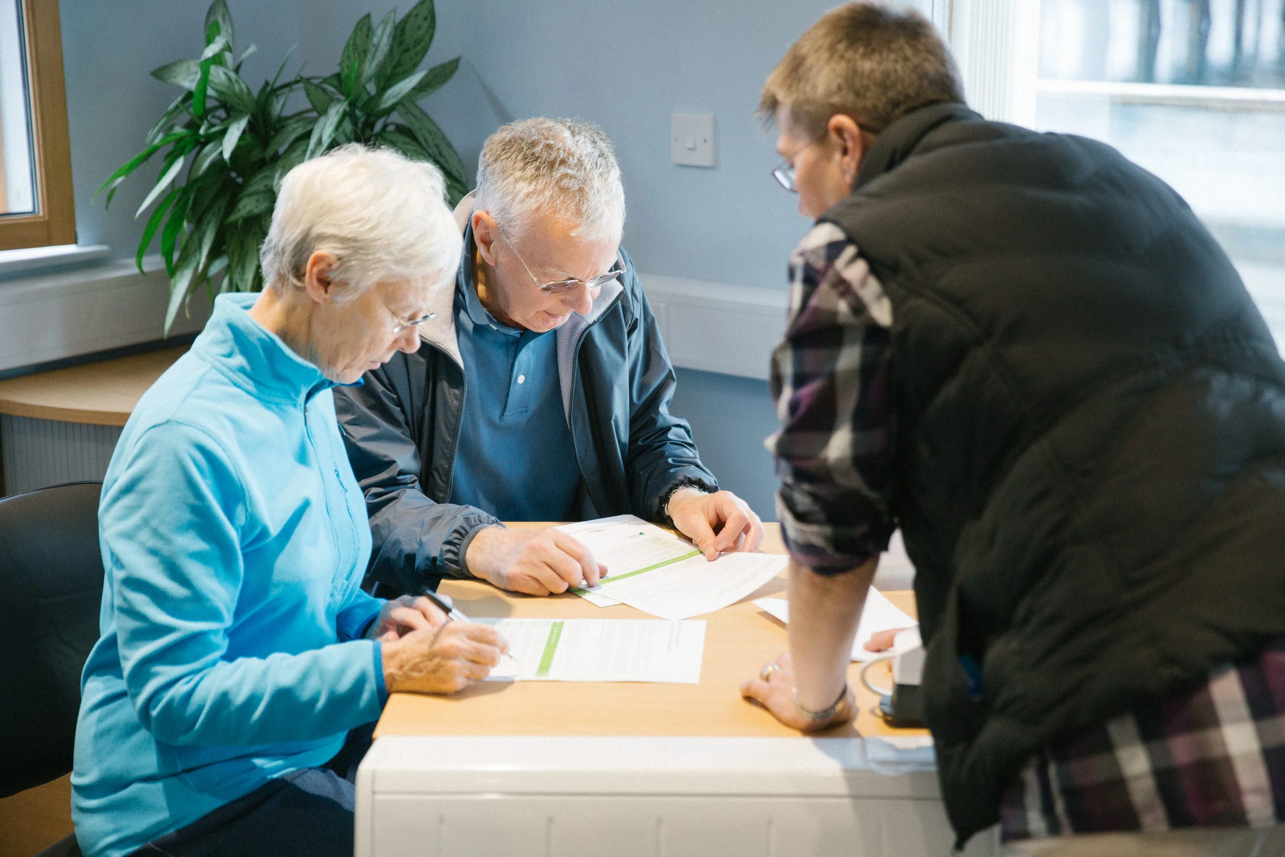 Three elderly people and a younger man gather around a table with papers and documents, engaged in a discussion or activity in a well-lit room with a window, green plant, and electrical outlet.