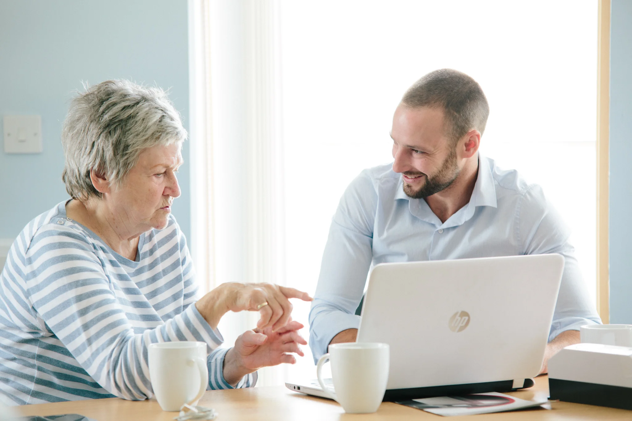 An elderly woman and a young man sit across from each other at a desk with a laptop, a computer mouse, and white coffee mugs. They are engaged in a conversation, with the woman pointing at the laptop screen and the man smiling.