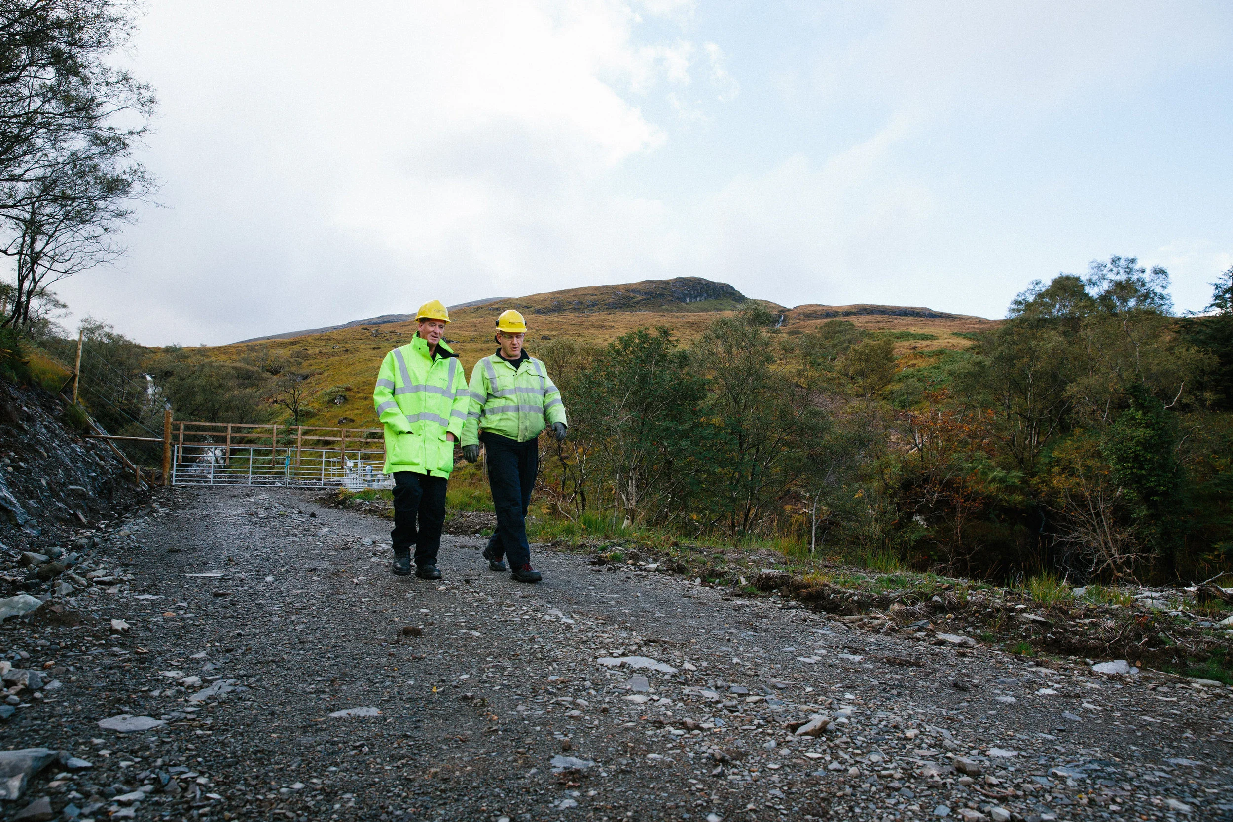 Two workers in yellow safety helmets and high-visibility jackets walking on a gravel path through a hilly outdoor landscape with trees and mountains in the background.