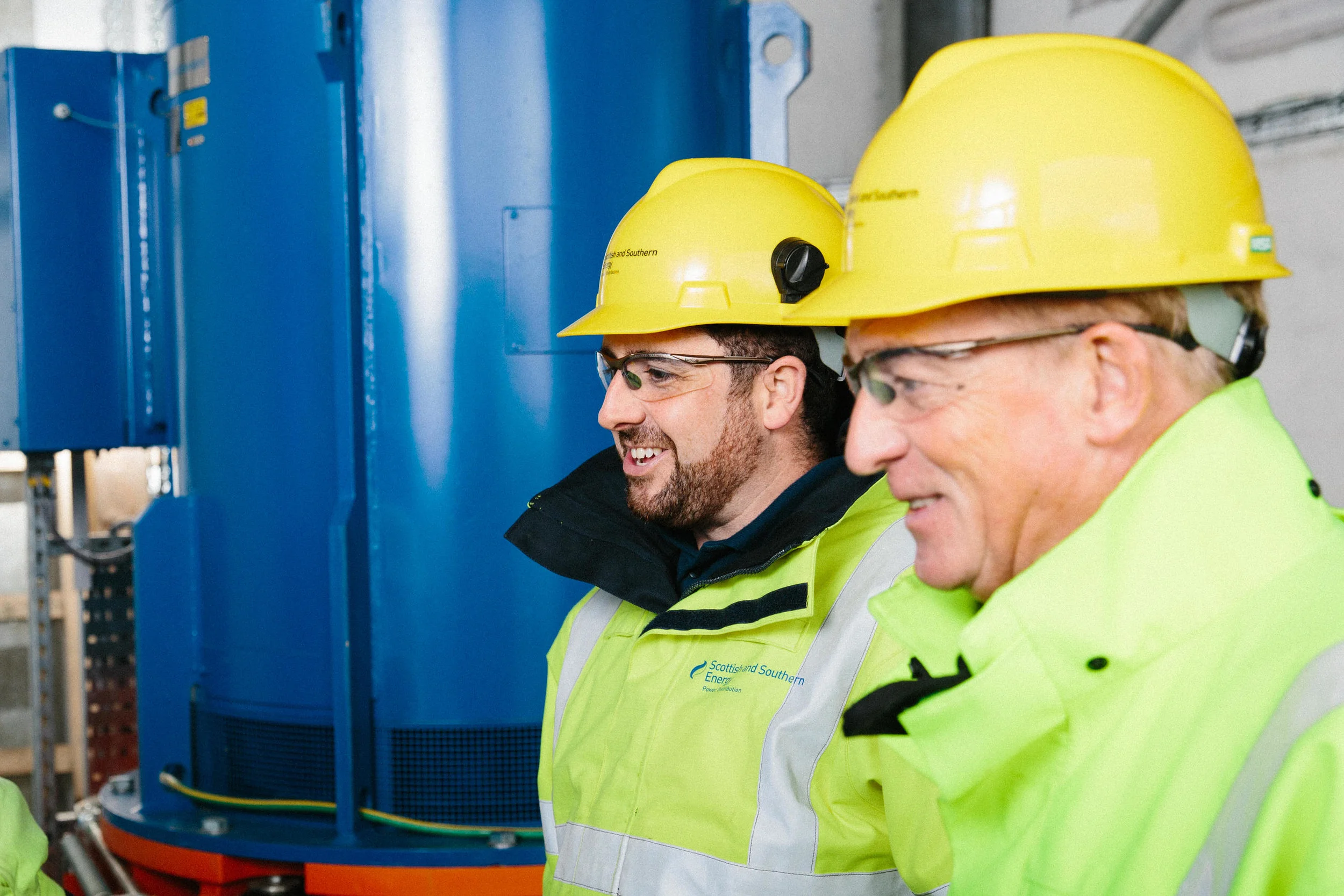 Two men wearing yellow hard hats and safety glasses working on blue industrial equipment in a factory or energy facility.
