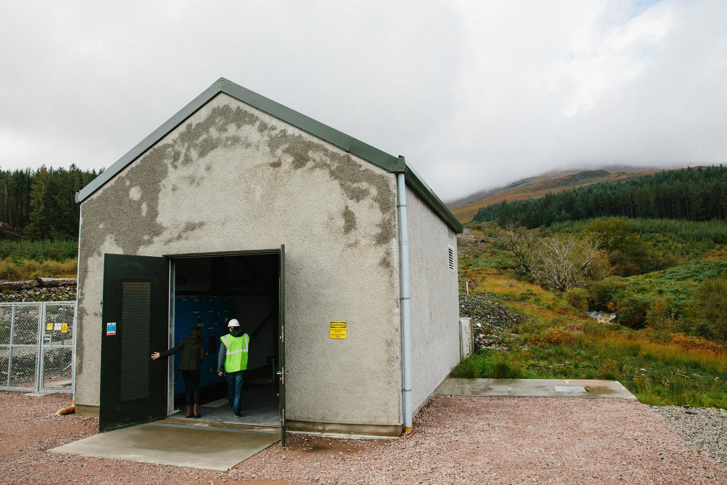 A small building with an open garage door, surrounded by a chain-link fence, with two people wearing hard hats and safety vests entering. The building appears weathered with patches on the exterior wall, set against a landscape with green trees and h