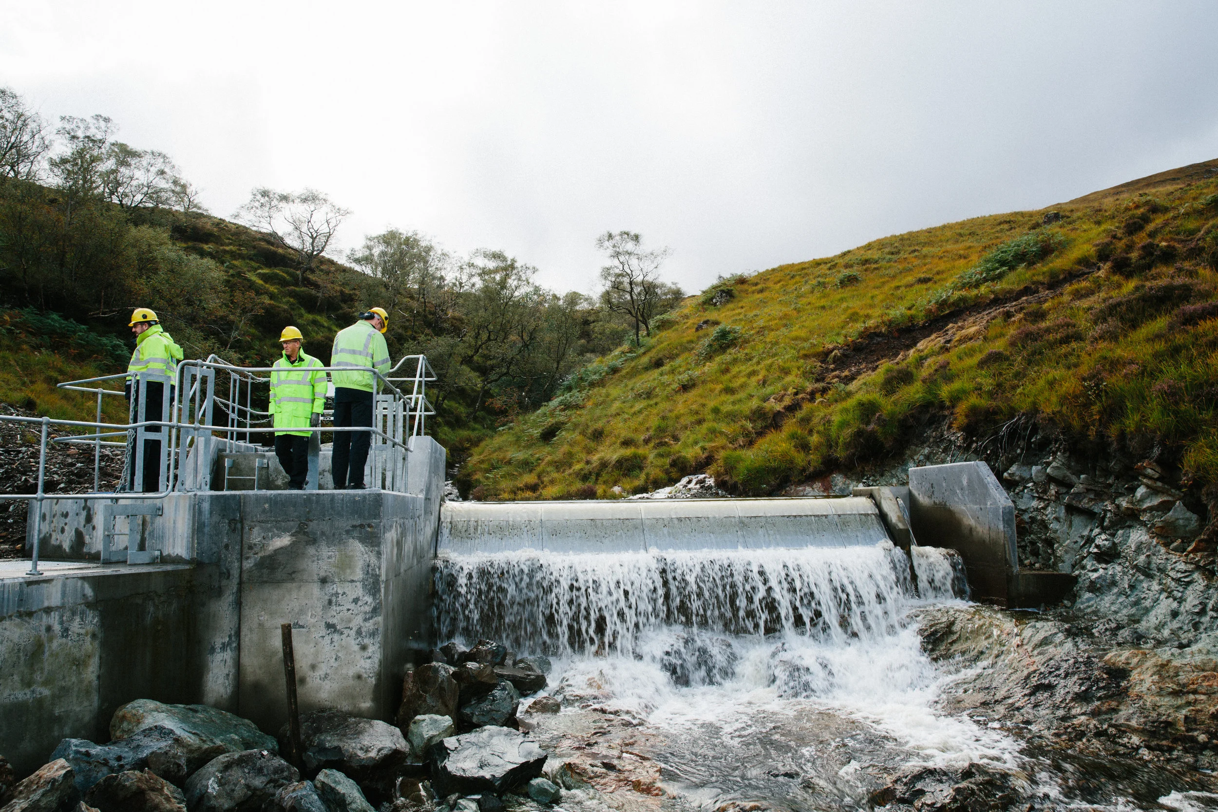 Three workers in yellow safety helmets and high-visibility jackets inspecting a dam with water flowing over it in a hilly, green landscape under a cloudy sky.