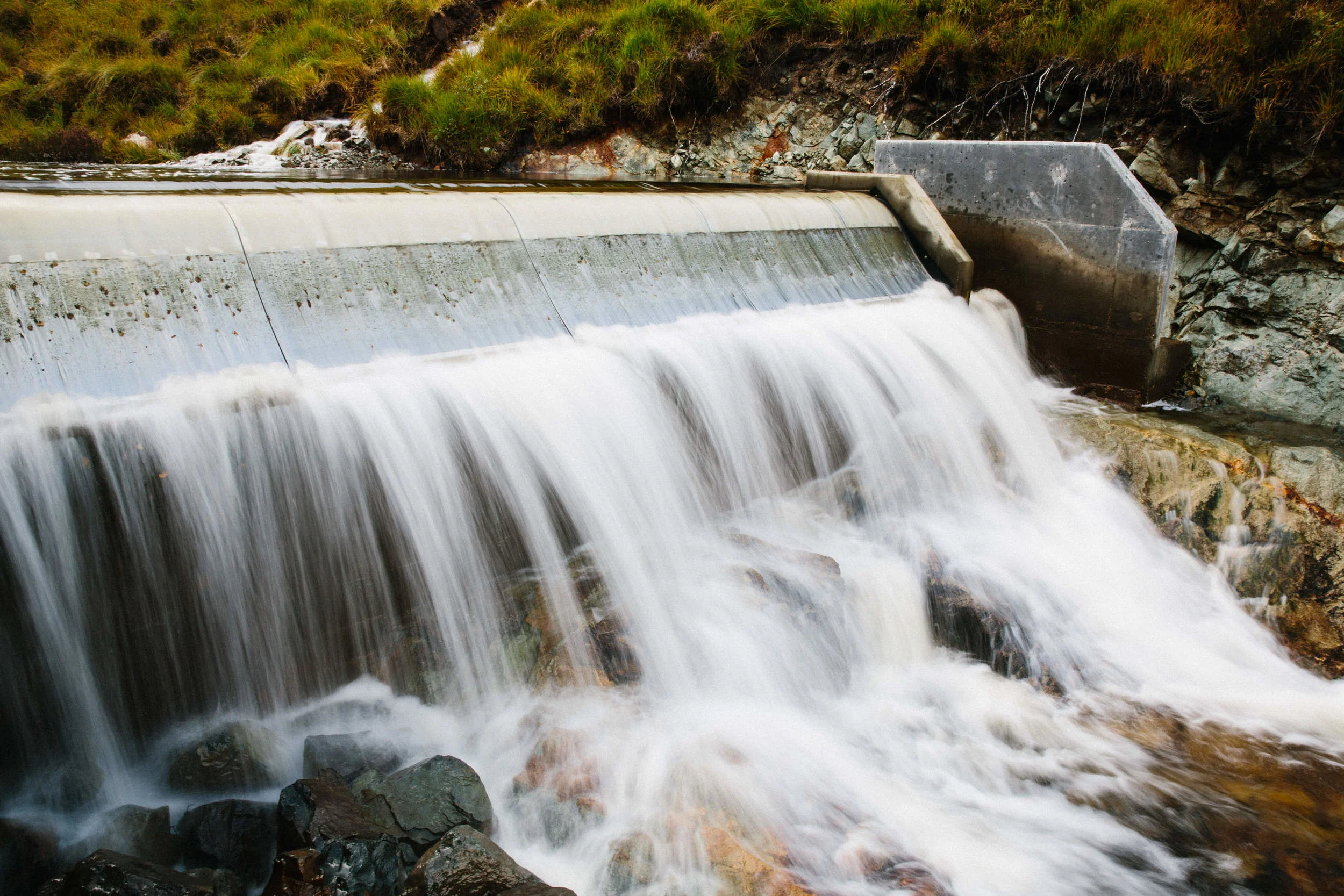 Water flowing over a dam with rocks at the base and grassy hillside above.