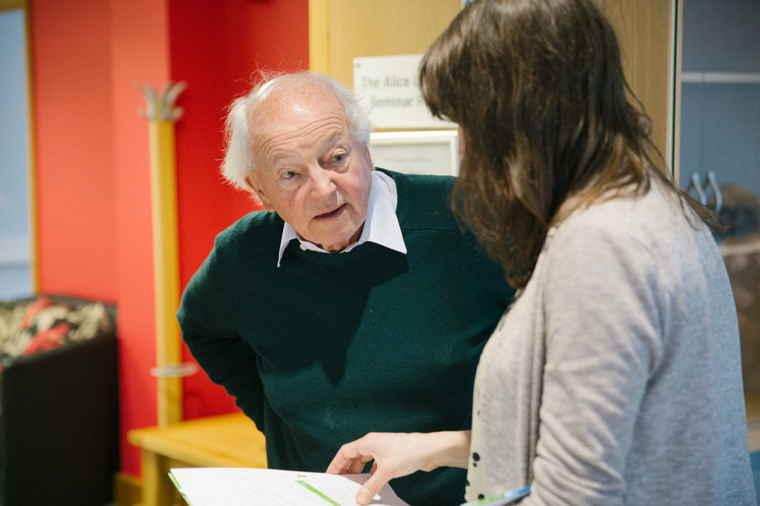 An elderly man with white hair talking to a woman in a gray sweater as she holds a booklet, in an indoor setting with colorful walls and a sign in the background.