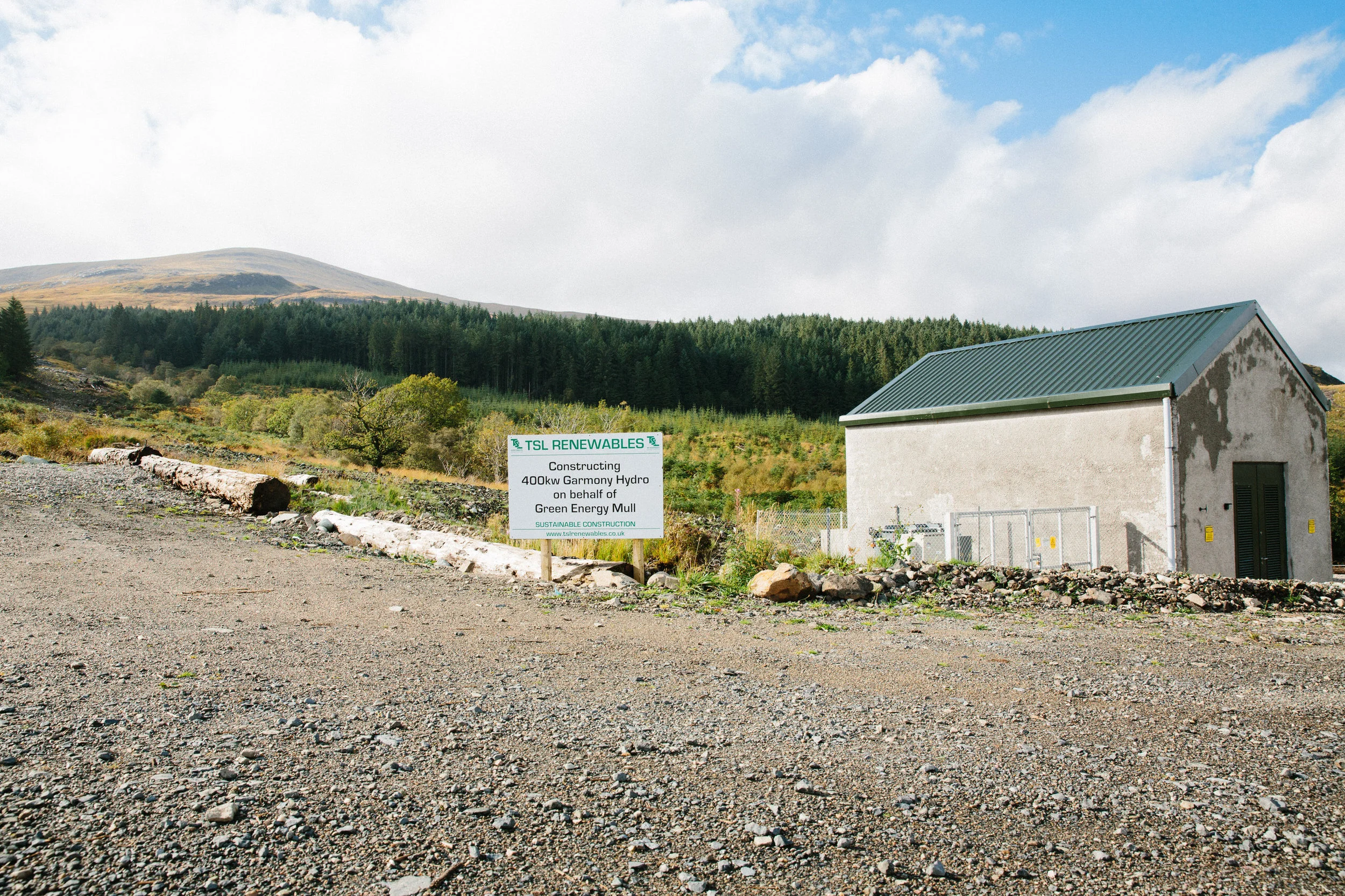 A rural scene with a gravel surface in the foreground, a small building with a green metal roof on the right, and a sign in front of the building that reads: 'TSL RENEWABLES - Constructing 400kw Garmony Hydro on behalf of Green Energy Mull.' In the b