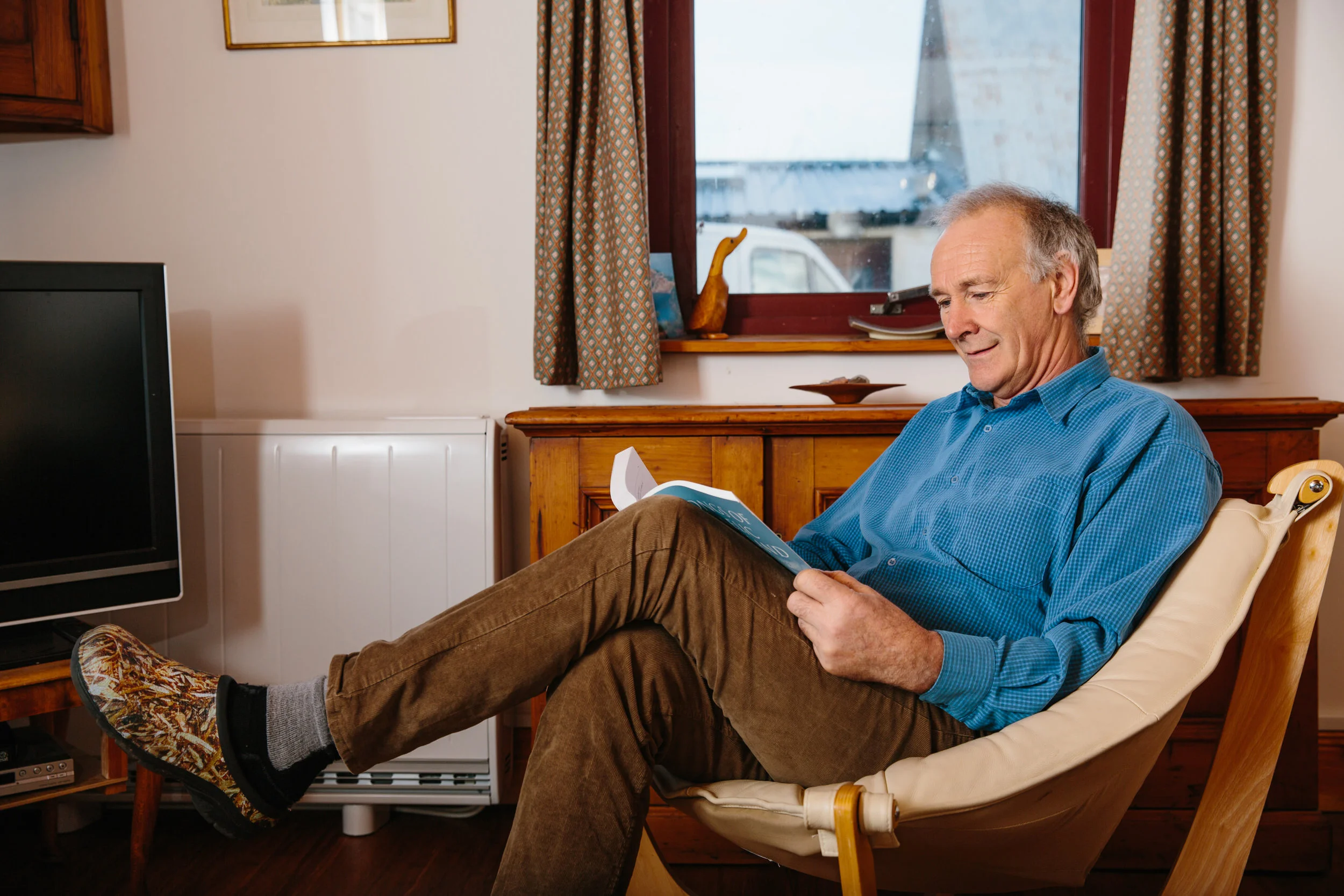 A senior man in a blue shirt and brown pants reading a book while sitting in a beige upholstered chair with his feet resting on a footstool. He is in a cozy room with a window, patterned curtains, a wooden side table, and a TV.