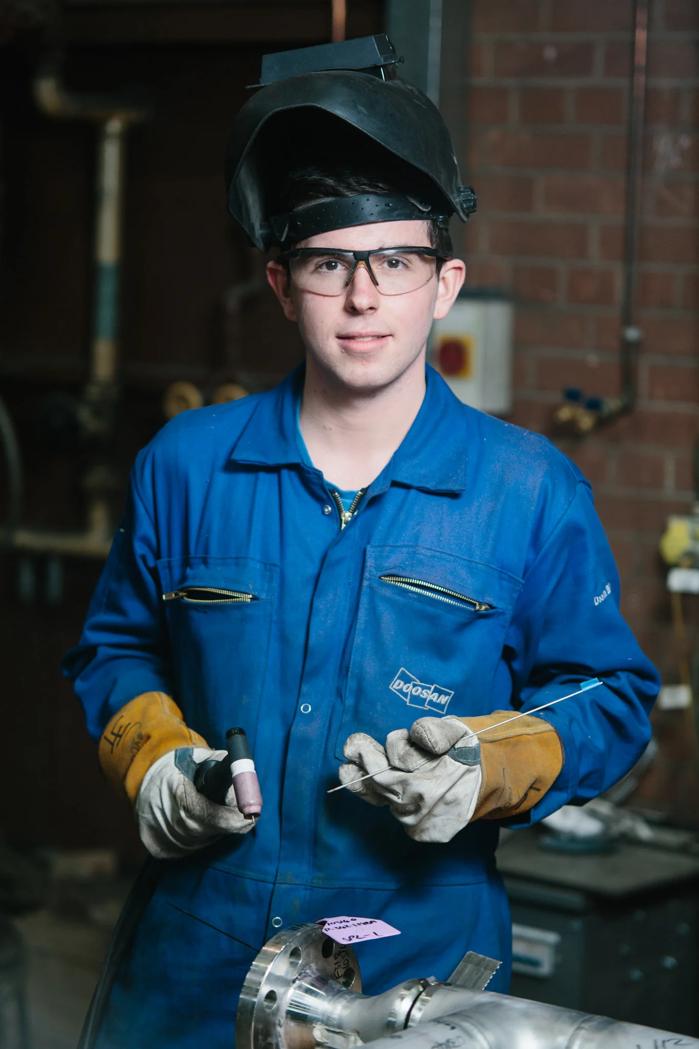 A man in safety glasses, a welding helmet pushed back, and a blue coverall is welding metal pipes with a welding torch. He is wearing protective gloves and standing in an industrial workshop with brick walls and machinery.