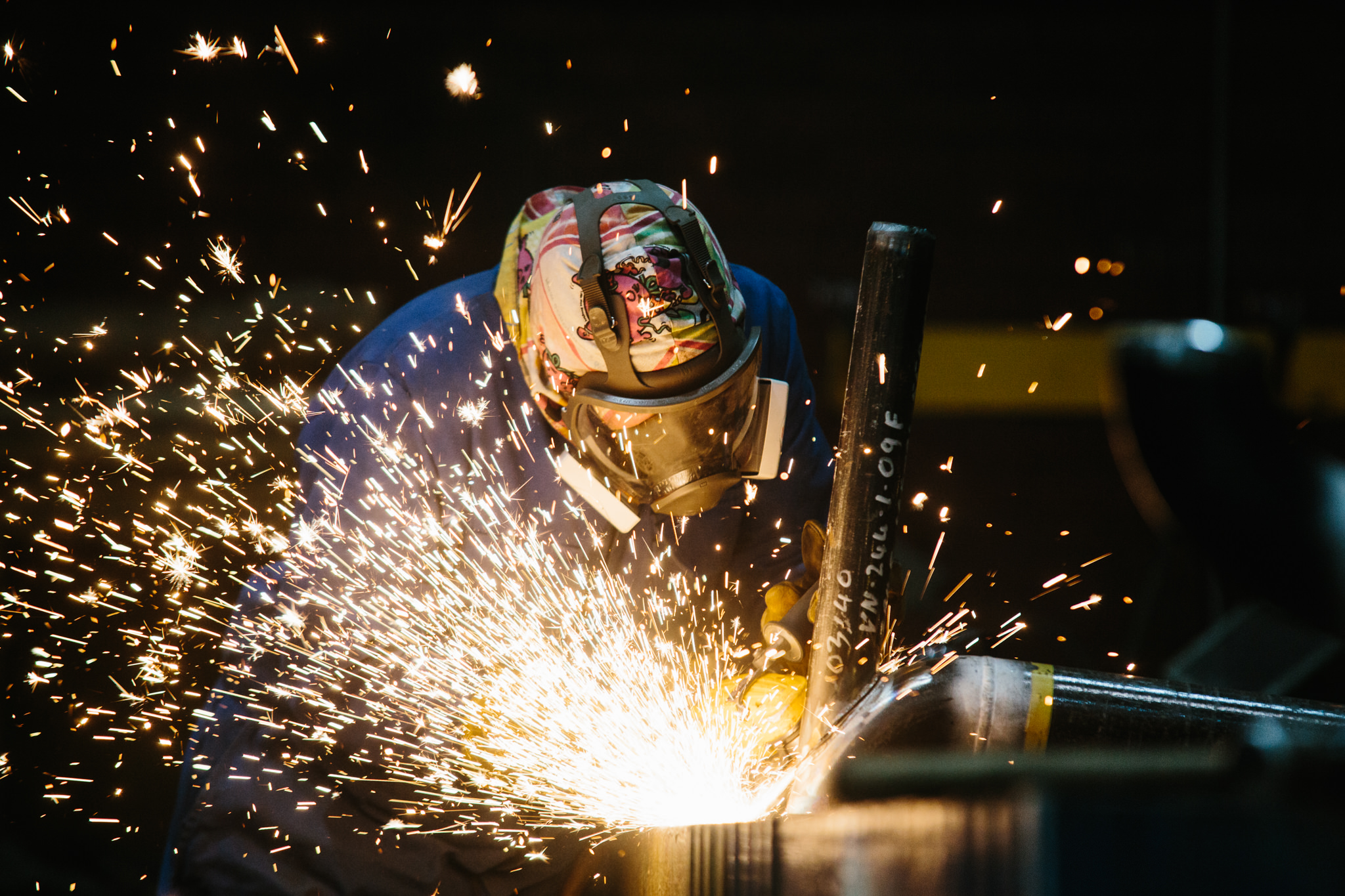 A worker wearing protective gear, including a helmet and goggles, welding metal with sparks flying around in a dark environment.