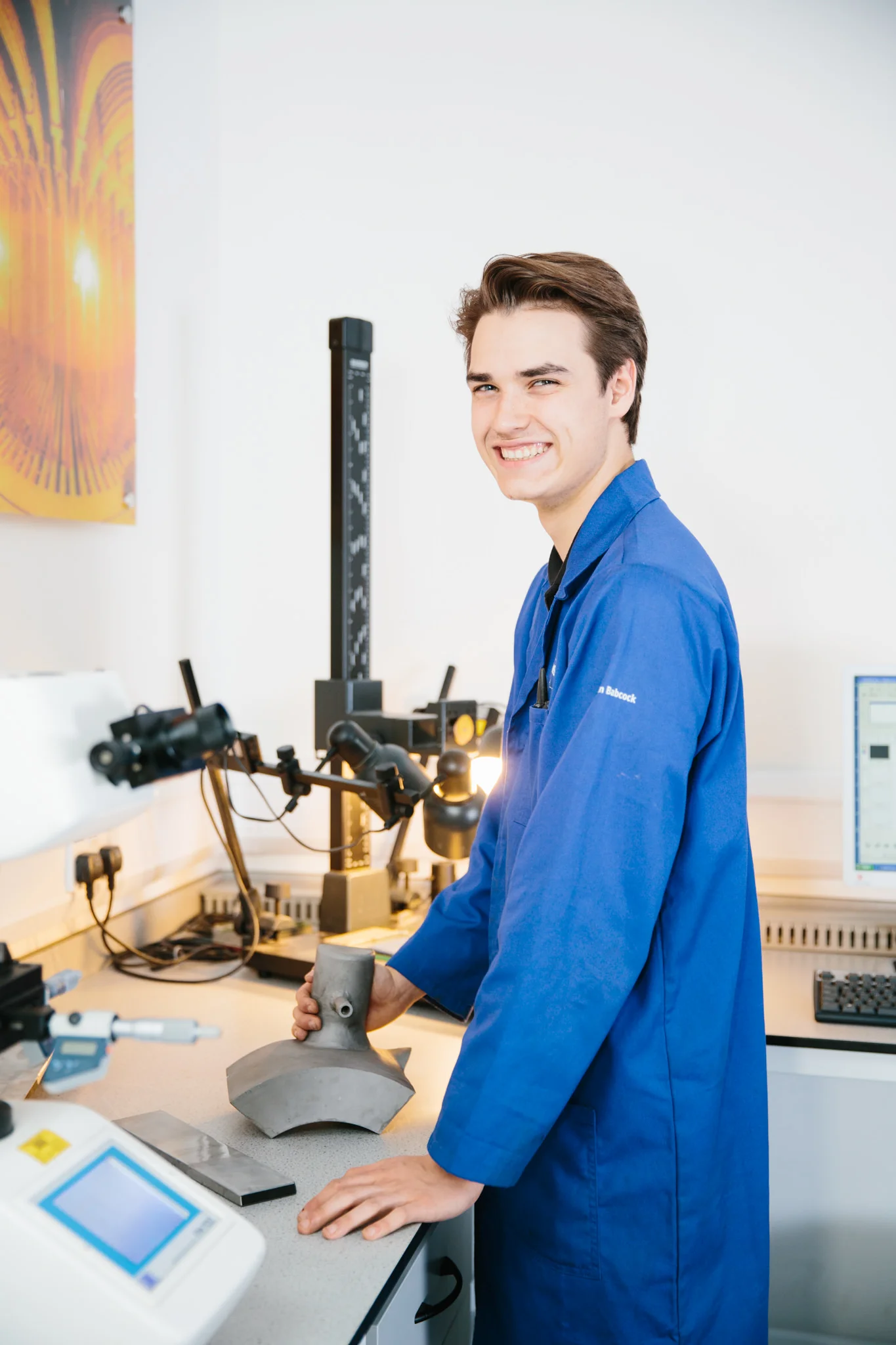 A young man in blue lab coat working in a laboratory, handling a metallic object, surrounded by scientific equipment including microscopes and computers.