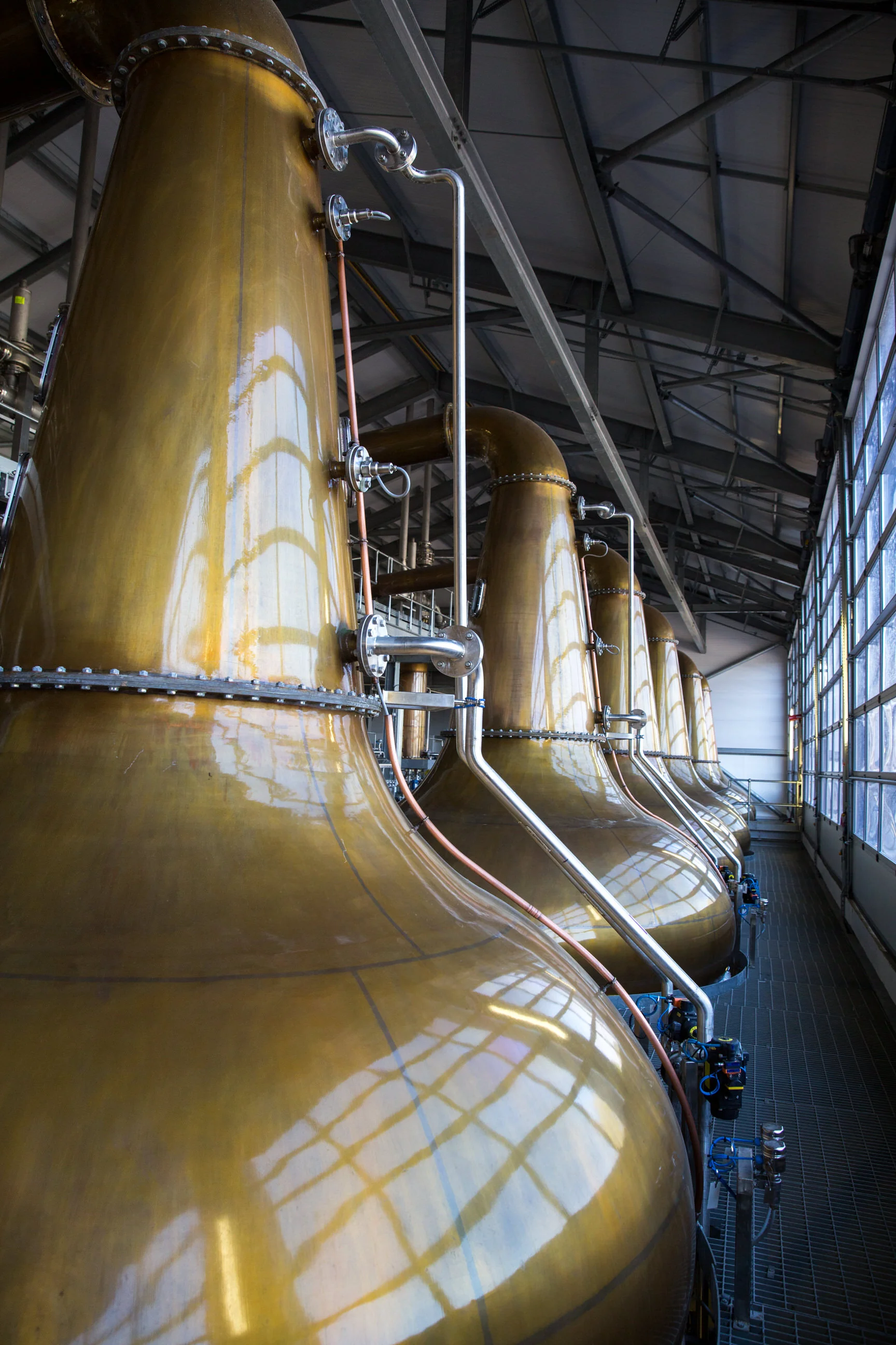 Large copper distillation tanks inside a distillery with industrial pipes and a high ceiling.