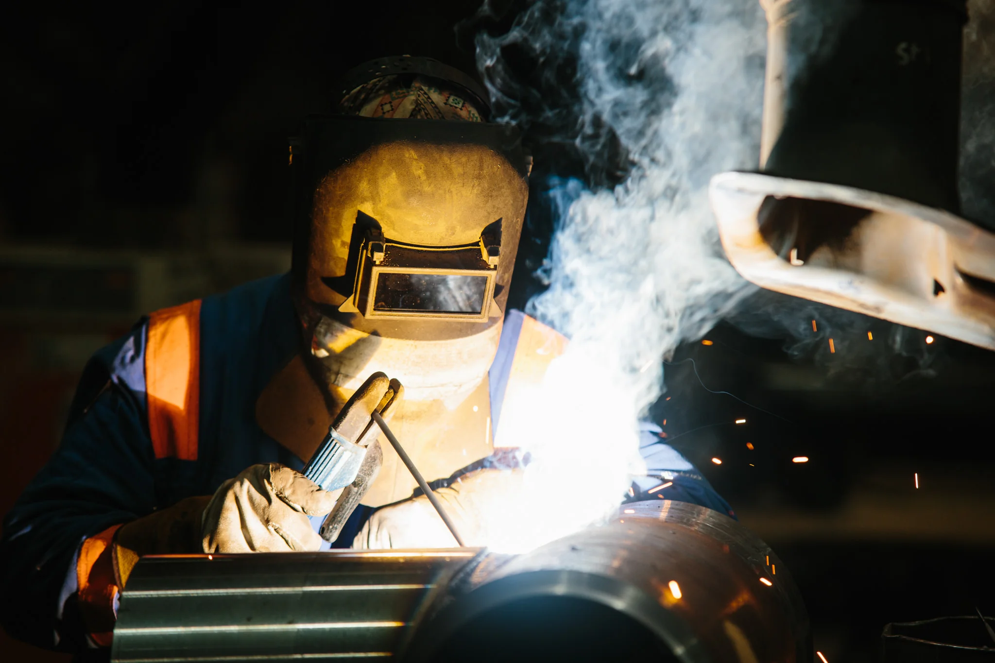 A worker welding metal pipe, wearing protective gear including helmet with face shield, gloves, and safety vest, with sparks and bright light from the welding process.