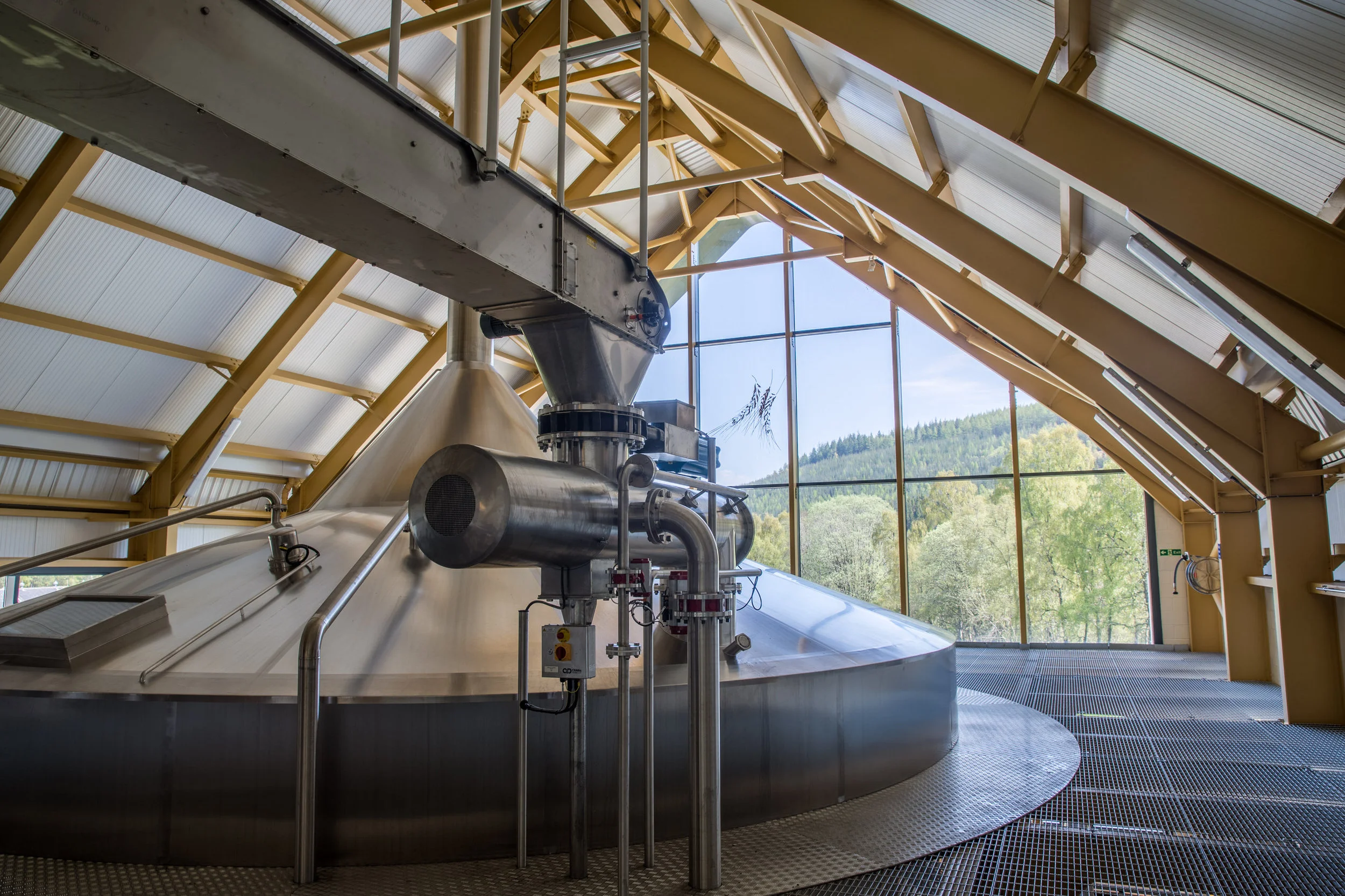 Interior of an industrial facility with large stainless steel machinery, pipes, and a sloped metal roof with large windows revealing a scenic outdoor view of trees and hills.