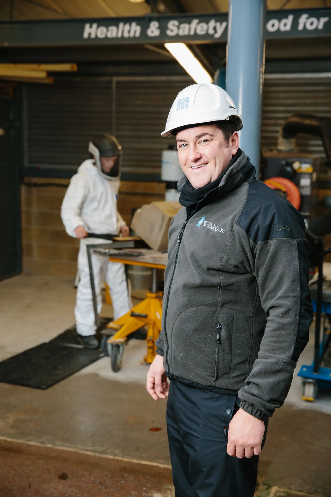 A man in a black jacket and pants wearing a white safety helmet standing in an industrial or workshop setting, with another person in white suit and black helmet working at a table in the background.