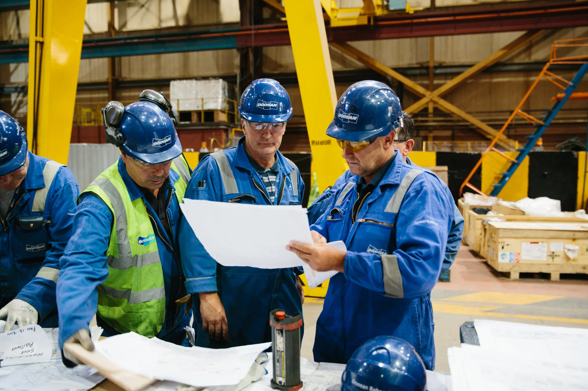 Group of construction workers wearing blue uniforms and helmets, examining documents in an industrial warehouse with equipment and crates in the background.