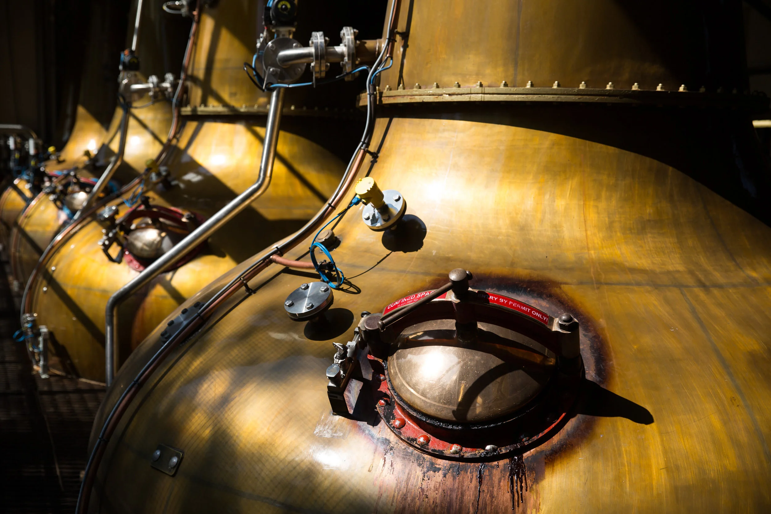 Close-up of an industrial tank with metal pipes and valves attached, with some wiring and a valve labeled 'COMMITTED SPACE' in red, and a black liquid pooled around the base of the valve.
