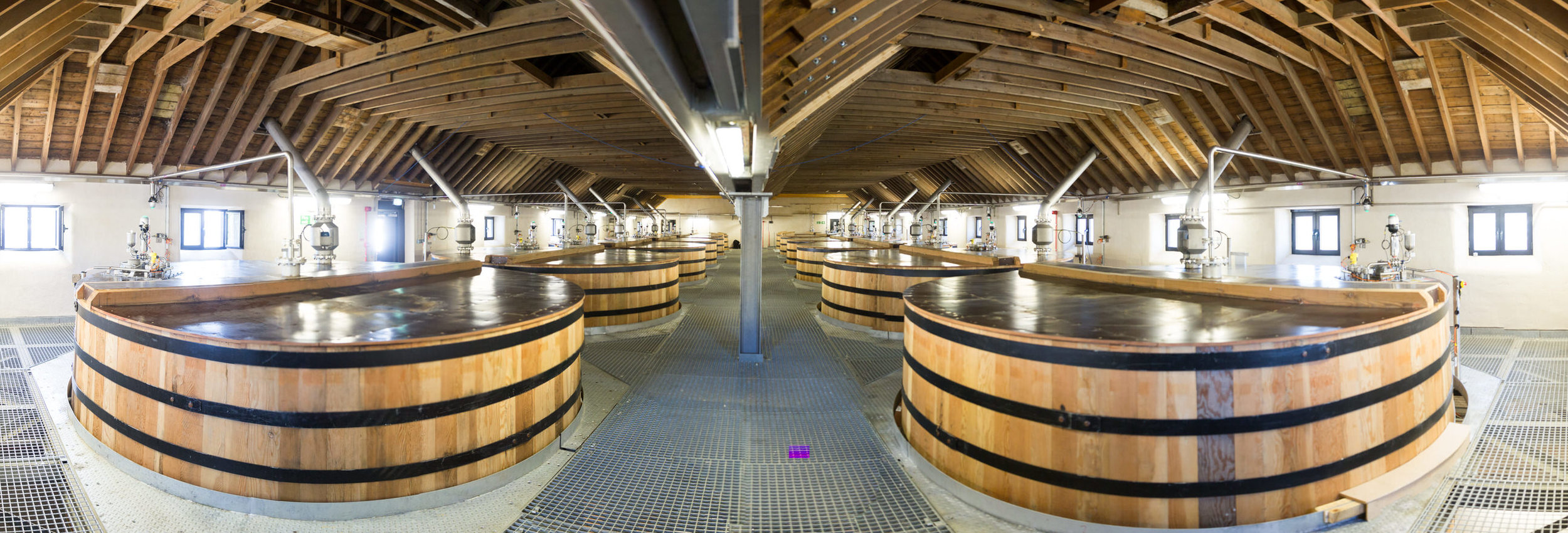 Empty large wine fermentation tanks in a winery’s fermentation room with wooden exteriors, located under a wooden ceiling with windows.
