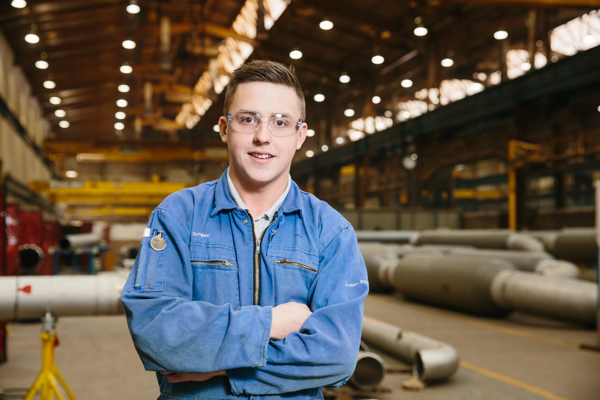 A young man in a blue work jacket and safety glasses stands with arms crossed inside an industrial facility with large pipes and overhead lighting.