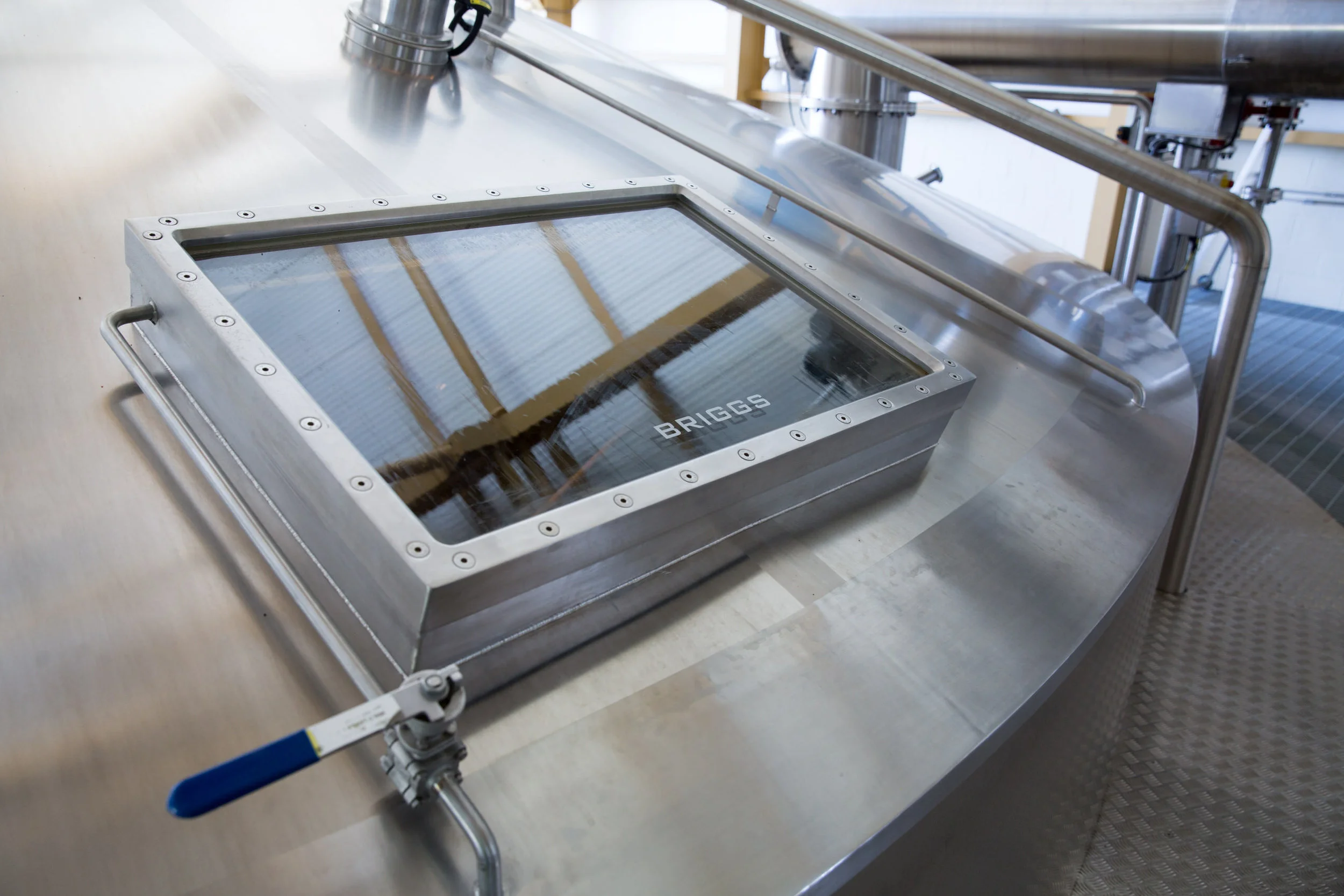 Stainless steel food processing equipment in an industrial setting, featuring a rectangular transparent container with the brand name 'Briggs' on it, attached to pipes and pipes with a blue handle.