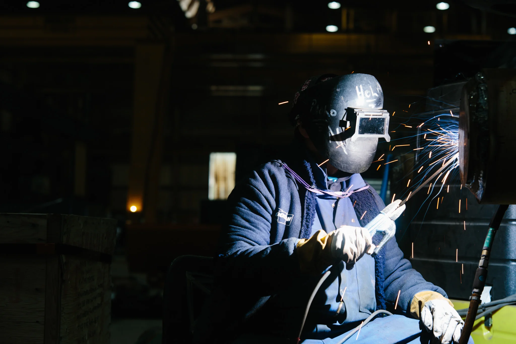 A person wearing a welding helmet and gloves welding metal, with sparks flying, in a dark workshop or industrial setting.