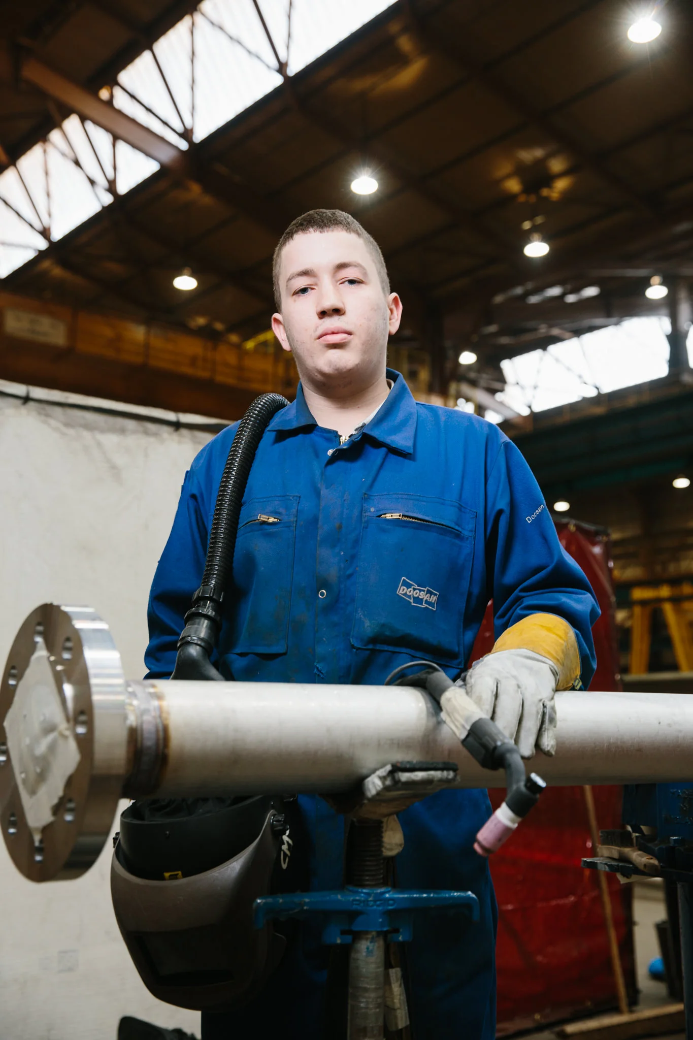 A young man in a blue work jumpsuit and gloves works with a pipe at a workshop or industrial setting, holding a welding or cutting tool.