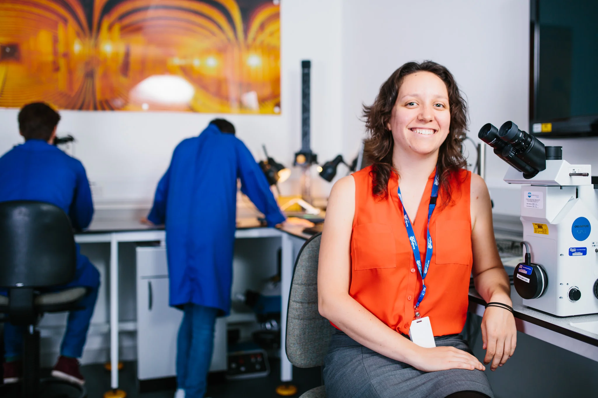 A woman with short curly hair smiling and sitting next to a microscope in a laboratory. In the background, two people are working at a desk with scientific equipment, wearing blue lab coats.