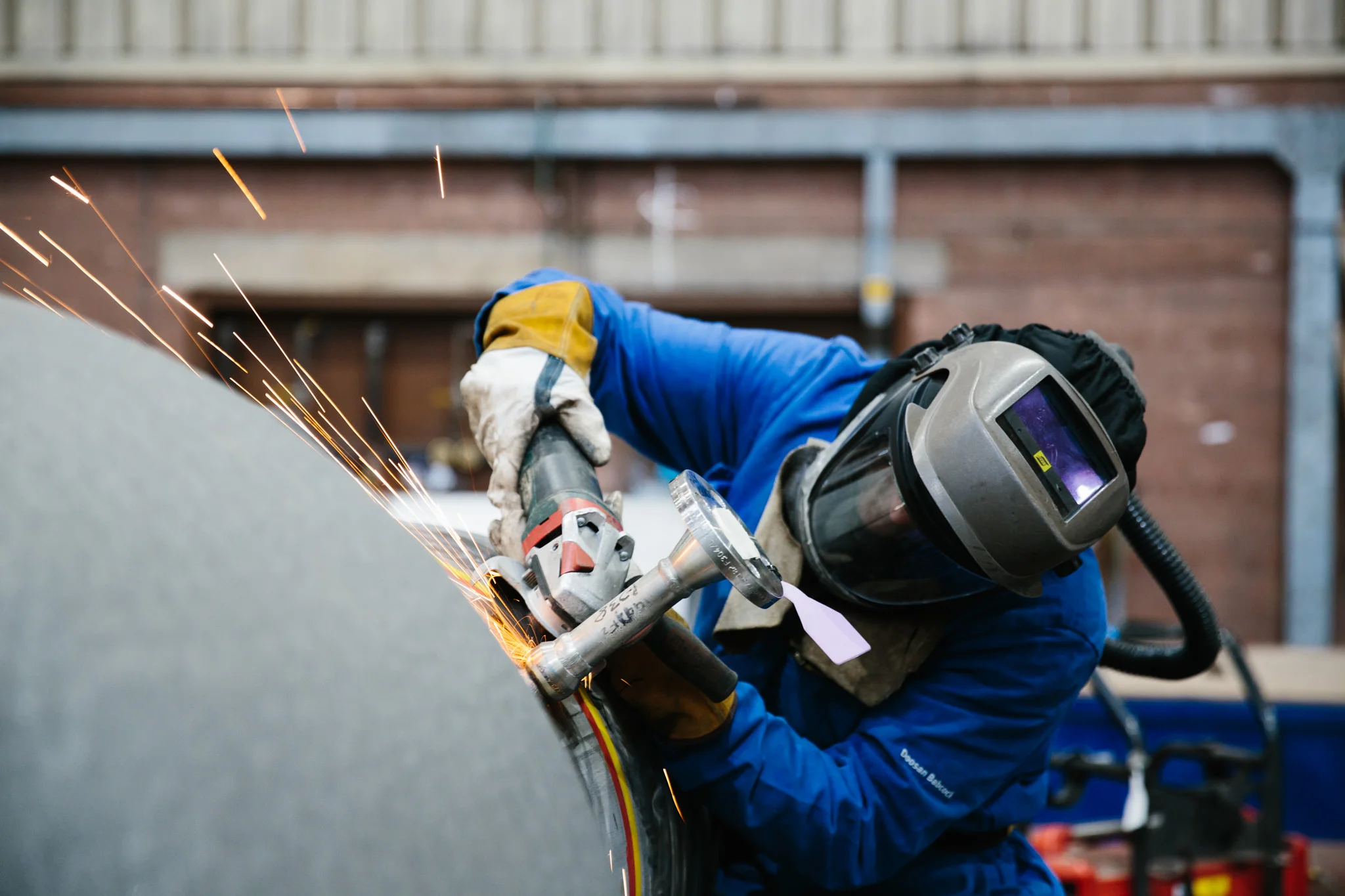 A construction worker wearing a welding helmet, gloves, and a blue jacket is welding a metal pipe, with sparks flying during the process, in an industrial setting.