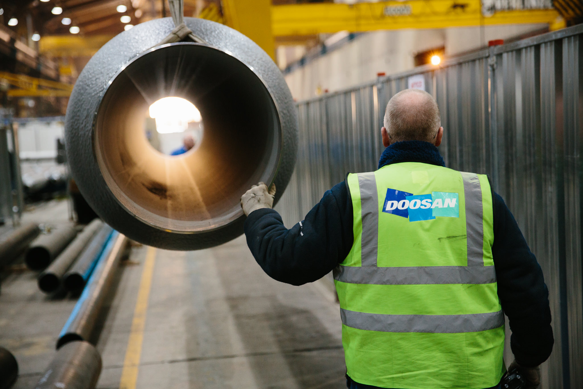 A worker in a neon yellow safety vest with the Doosan logo, looking at a large metal pipe inside an industrial warehouse.