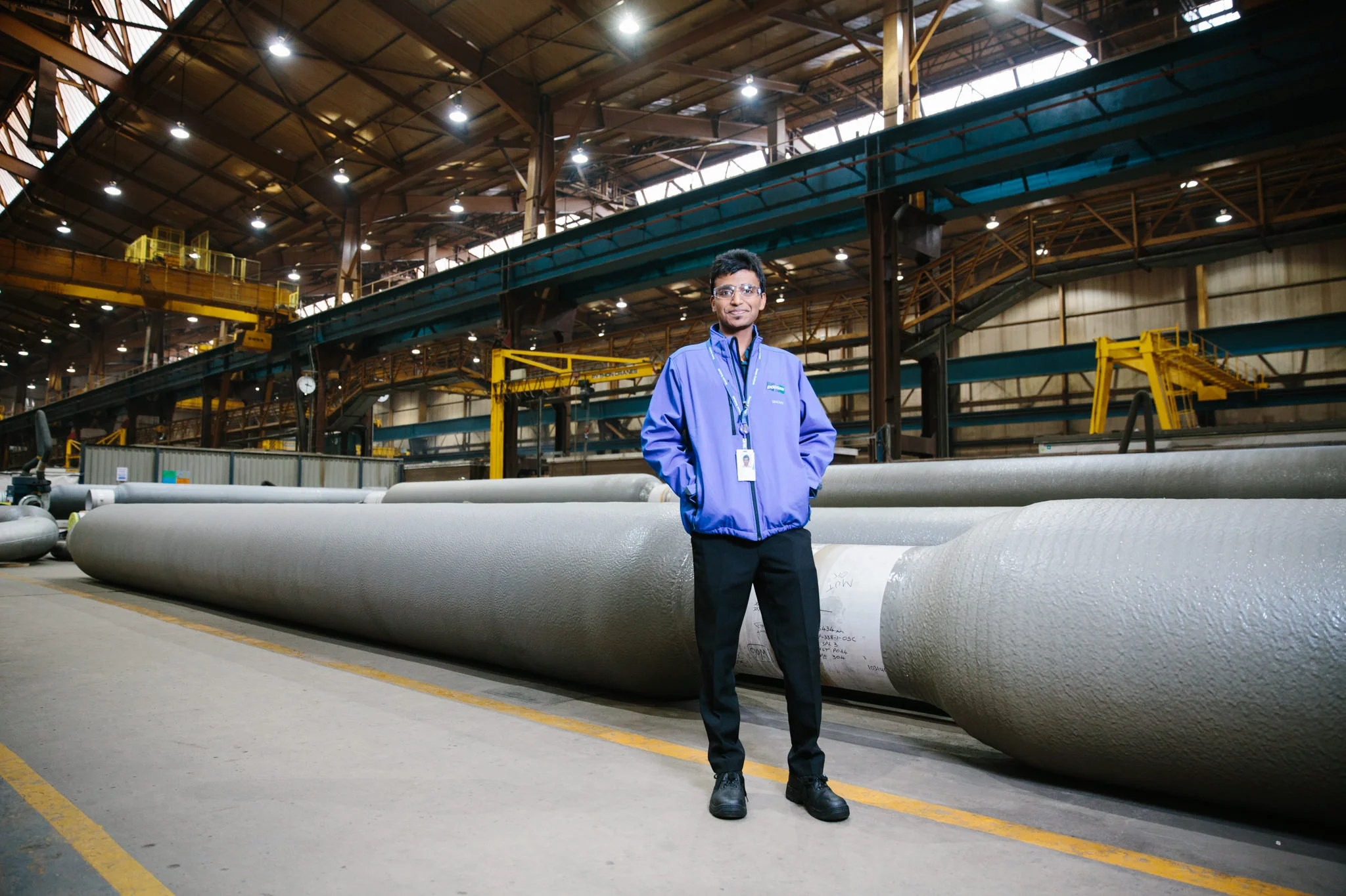 A man in safety glasses and a blue jacket stands in an industrial warehouse with large pipes on the floor. The warehouse has a high ceiling with metal beams, overhead lighting, and yellow overhead cranes.