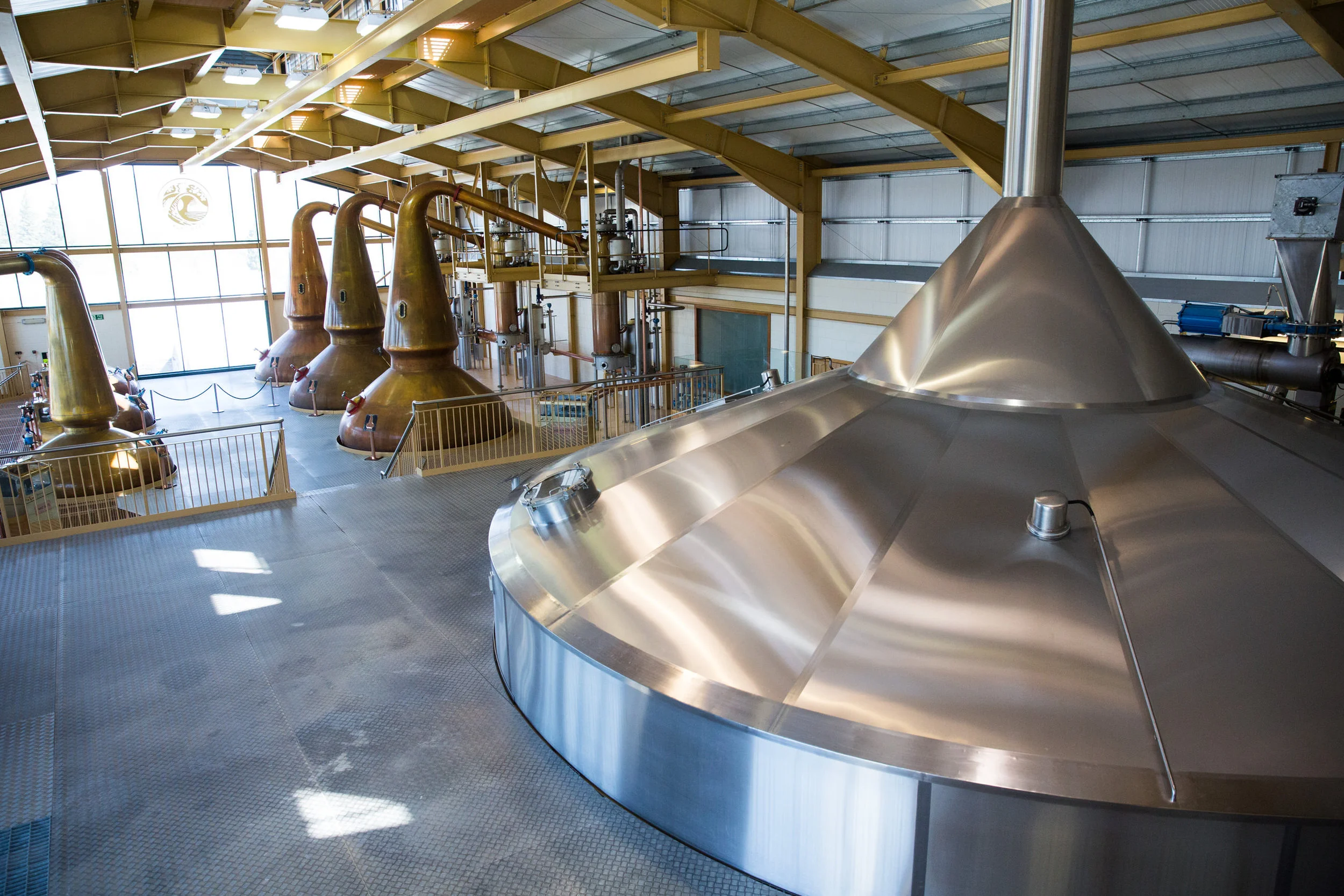 Inside a distillery with large copper stills and stainless steel equipment, bright lighting, and large windows.