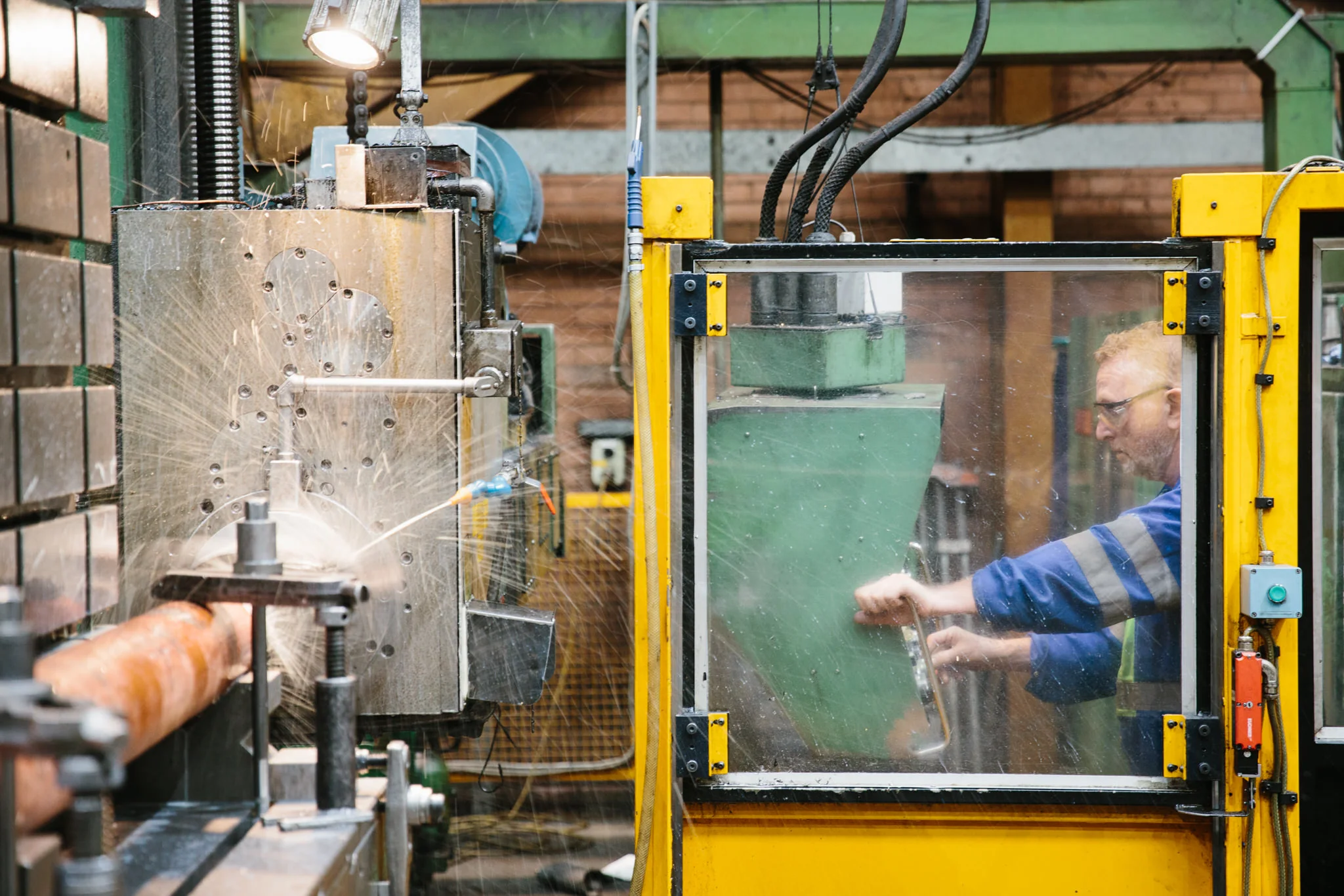 A man with safety glasses operating machinery inside a factory, with metal parts and sparks flying from the equipment.